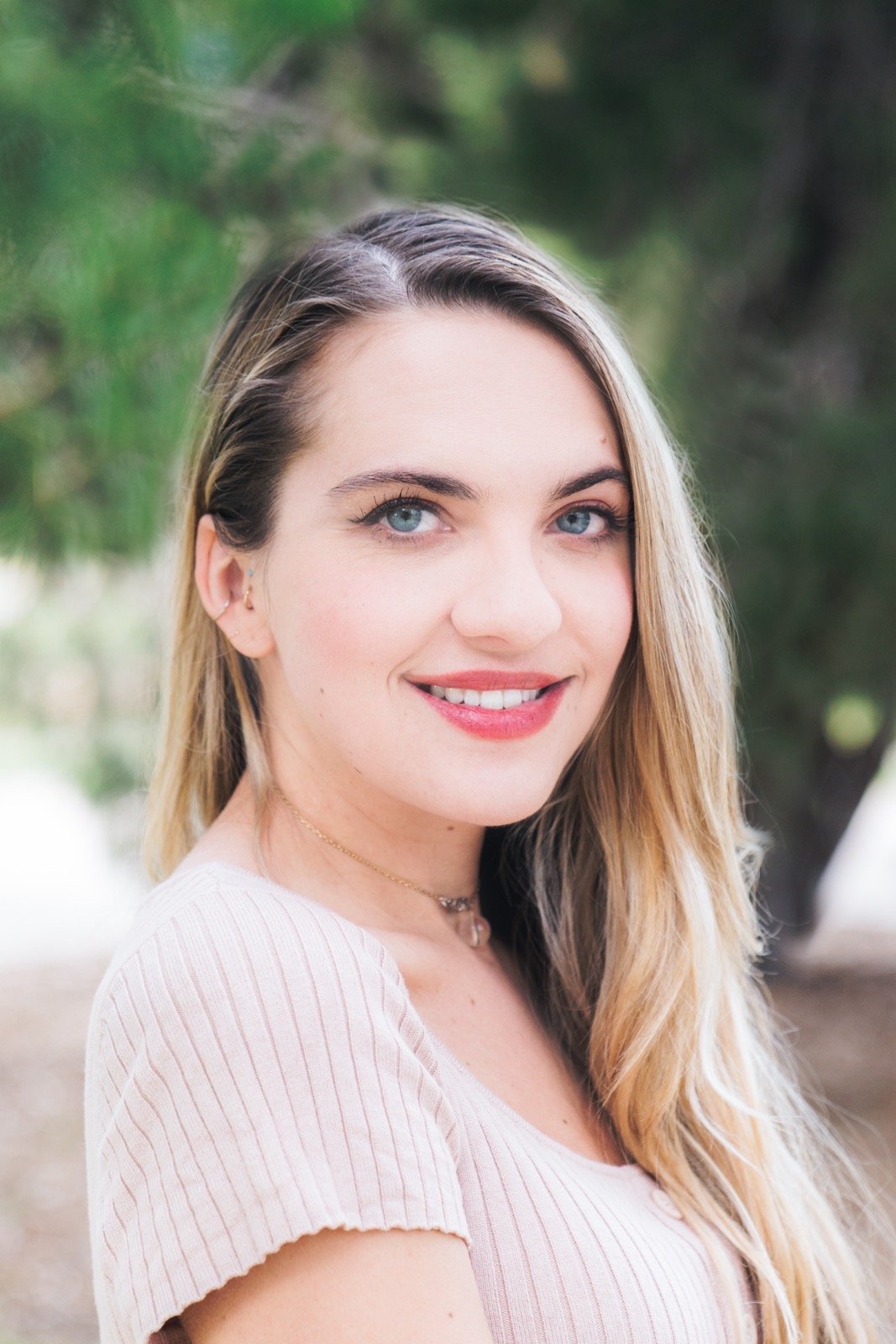 Smiling woman with long blond hair wearing a pink top, standing outdoors with green foliage in the background.