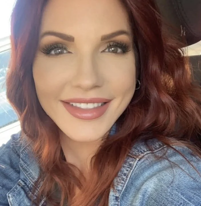 Woman with wavy red hair, wearing a denim jacket, smiling.