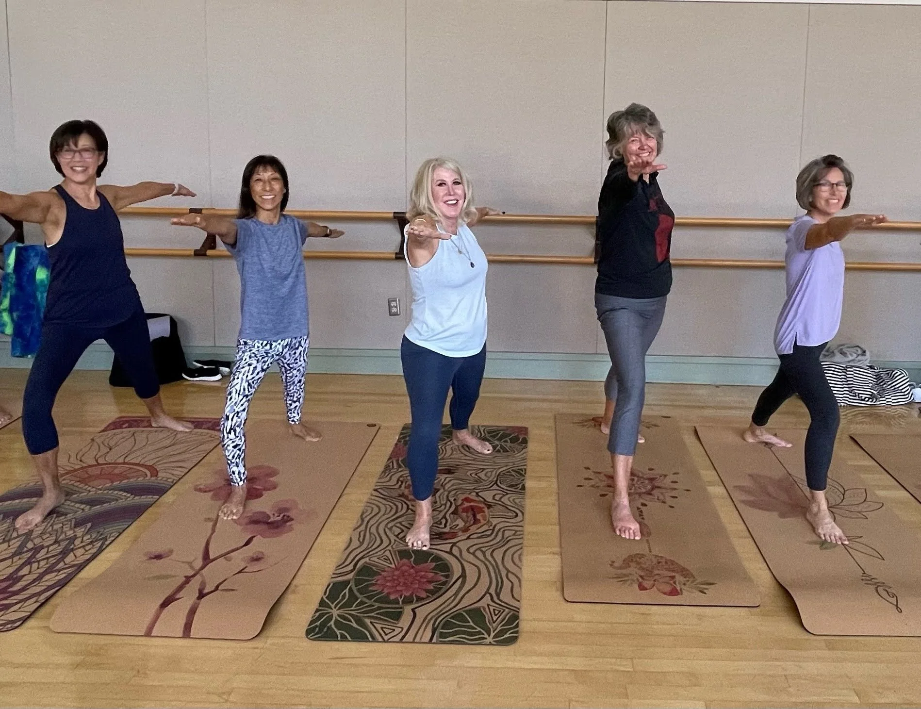 Five women participating in a yoga class, standing on mats in a dance studio, reaching out with one arm extended, smiling, with a ballet barre behind them.