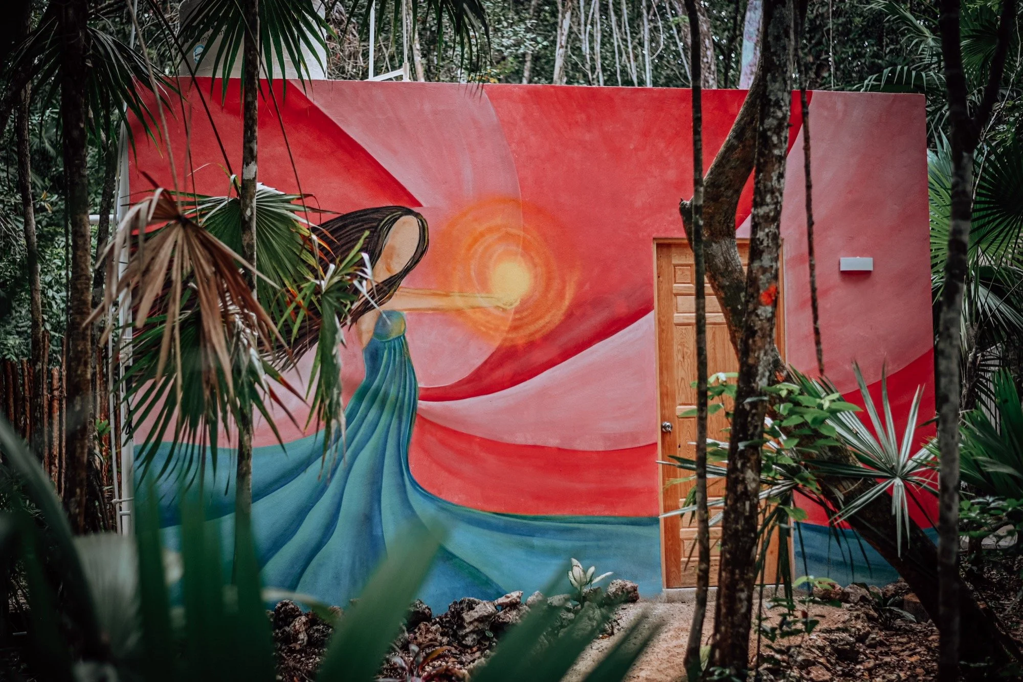 Colorful mural of a woman with long dark hair in a flowing blue dress, with a glowing orange sun in the background, painted on an outdoor wall surrounded by green tropical plants.