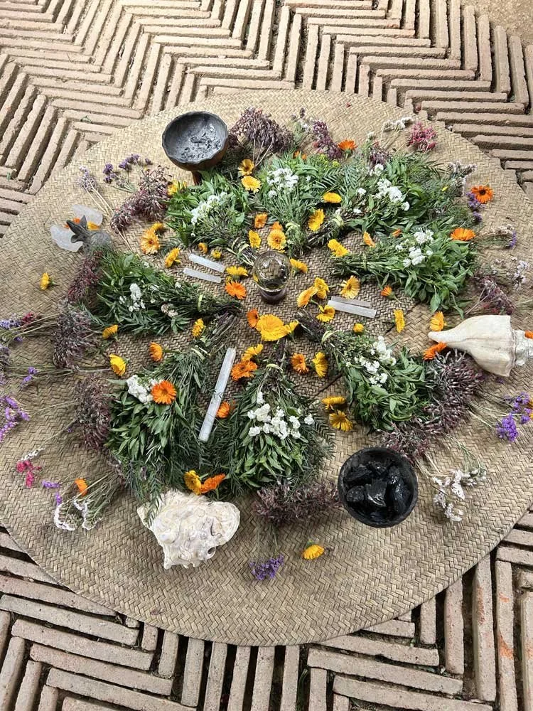 A circular arrangement of various flowers, leaves, and natural objects laid out on a woven circular mat, with small bowls around it containing candles and stones, on a brick surface.
