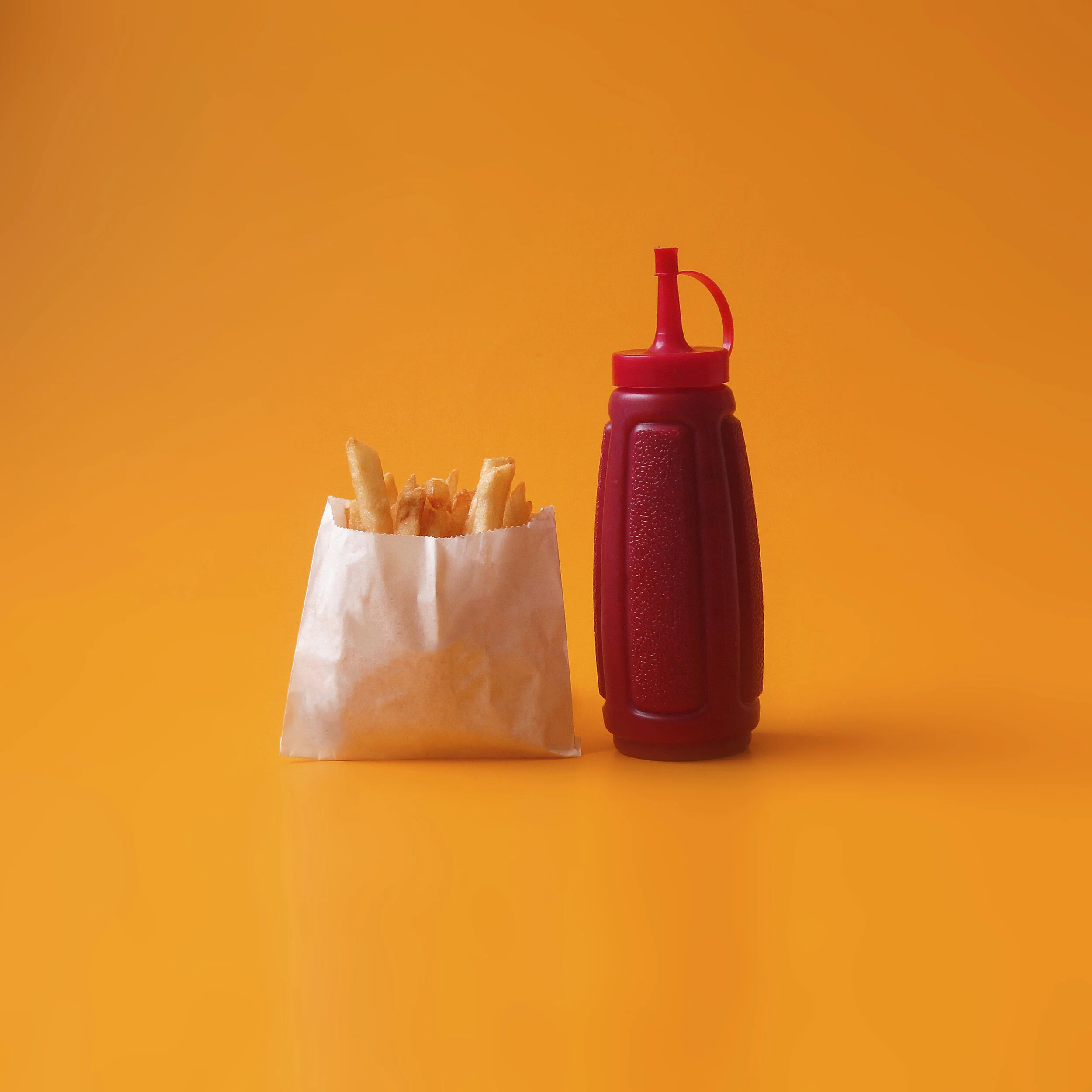 A paper bag of French fries next to a red squeeze bottle of ketchup on an orange background.