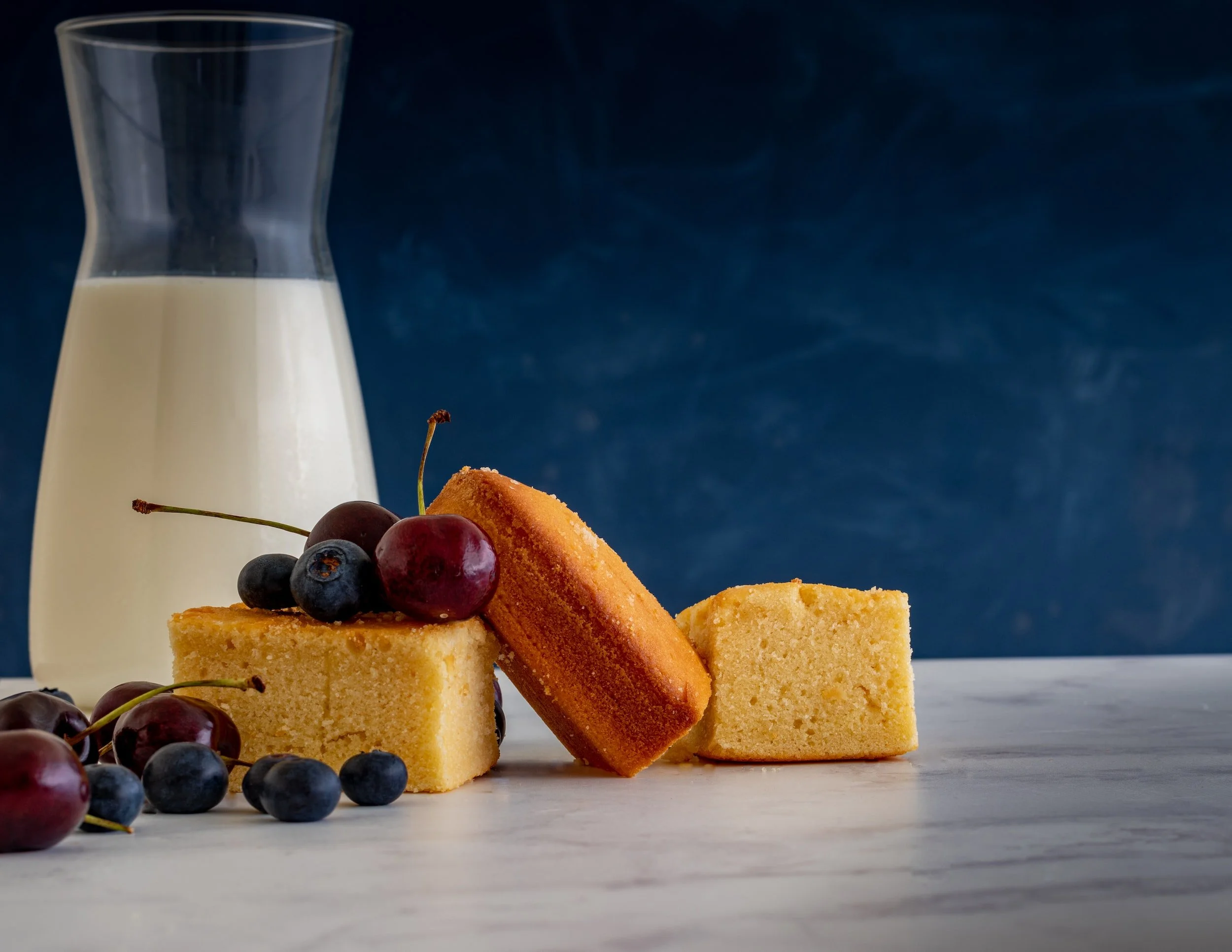 Slices of golden cake arranged with fresh cherries and blueberries on a marble surface, with a glass pitcher of milk in the background.