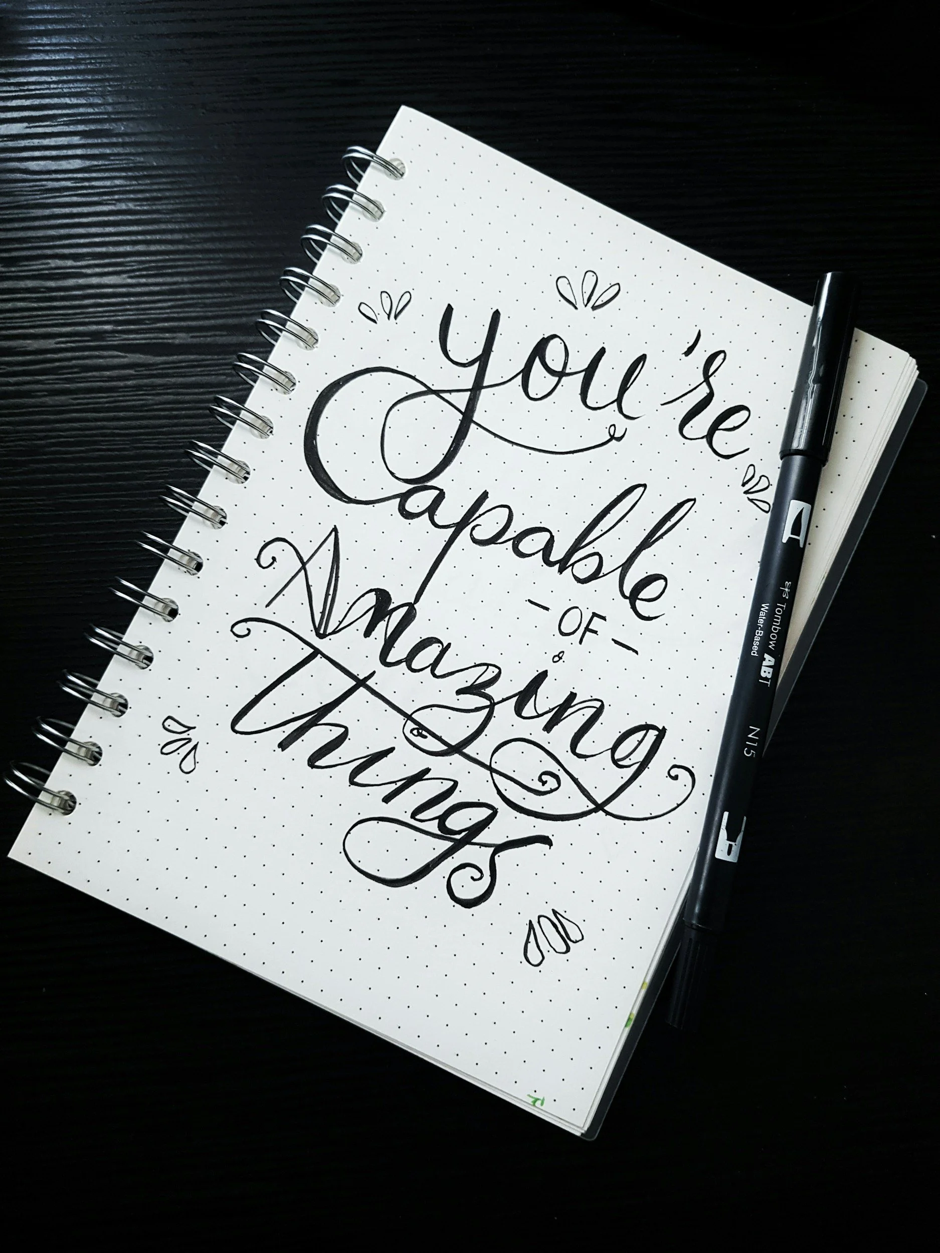Open notebook with hand-lettered quote “You’re capable of amazing things” written in elegant black script, with a black brush pen resting beside it on a dark wooden surface.