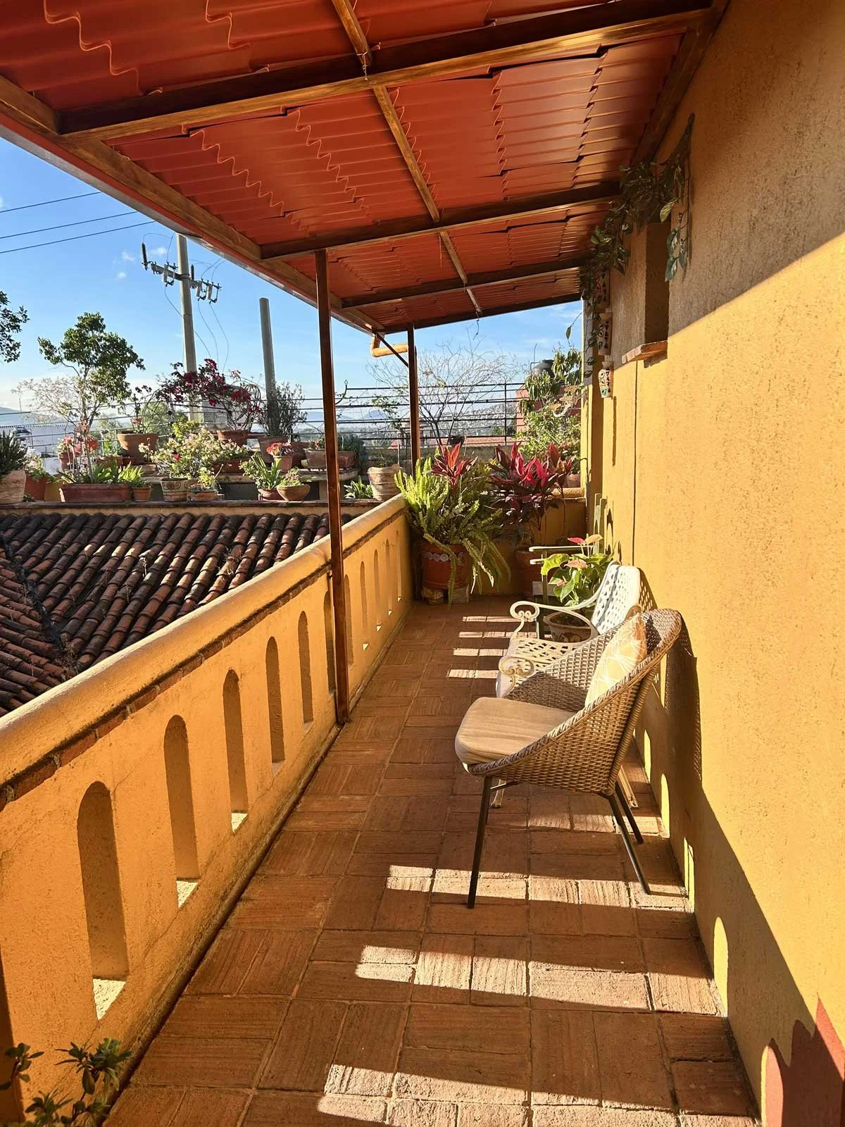 Sunlit balcony with yellow walls, terracotta tile flooring, potted plants, and wicker chairs, under a red corrugated roof.