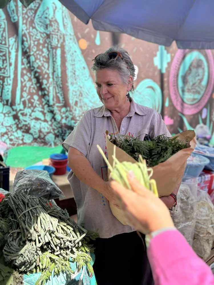 A woman at an outdoor market is holding a bouquet of fresh greens wrapped in brown paper, smiling as she receives it from a customer. The market stall is filled with various fresh vegetables, and a colorful, artistically decorated background is visib
