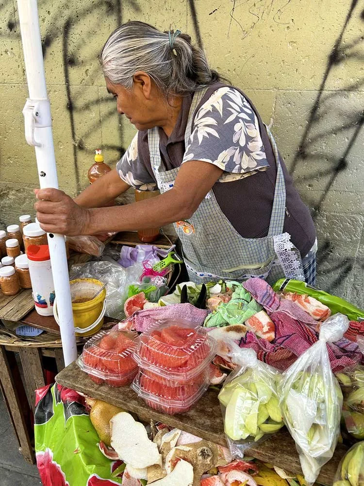 An elderly woman with gray hair, wearing a floral shirt and apron, is preparing food at her street market stall. The stall has cut watermelon, packaged vegetables, and various jars of spices or condiments on a wooden table.