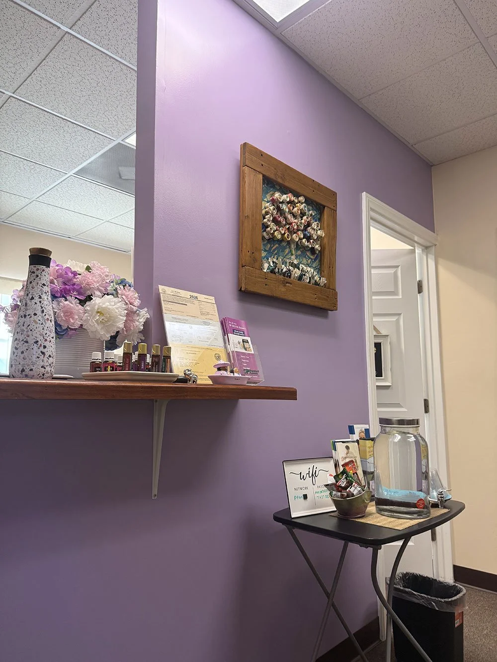Interior of a room with a purple wall, a wooden shelf holding decorative items and nail polish bottles, a framed art piece made of rolled nails, and a small black table with a glass jar of water, a bowl of snack chocolates, and some informational pamphlets.