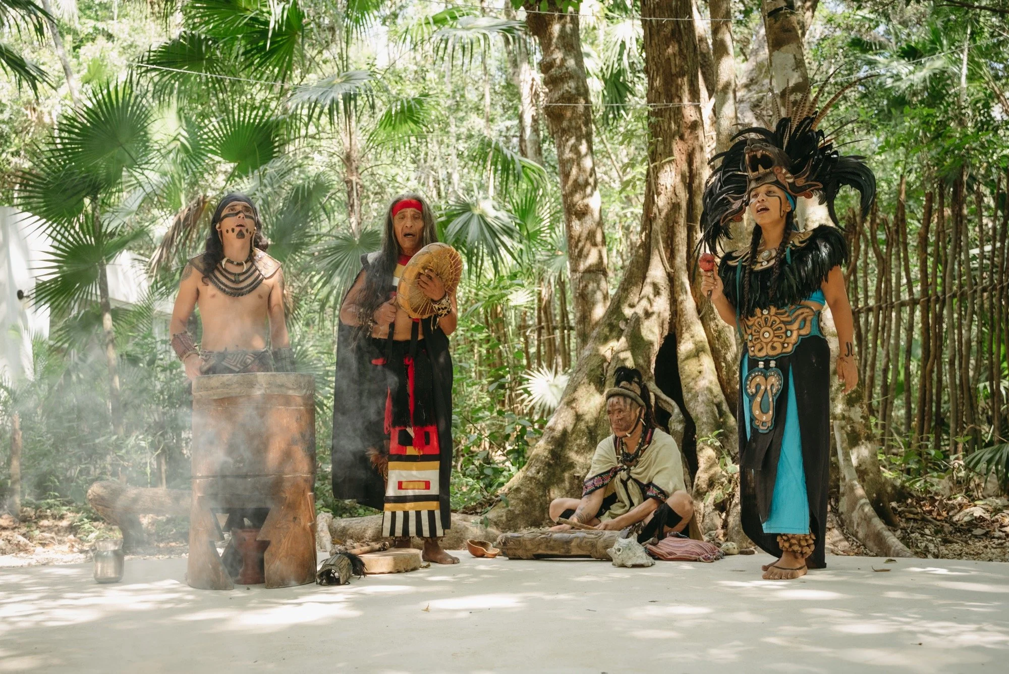 Five individuals in traditional indigenous attire performing a cultural ritual in a lush forest. One person is sitting on the ground, four are standing, one holding a drum, and another holding an ice cream cone. They are surrounded by green tropical