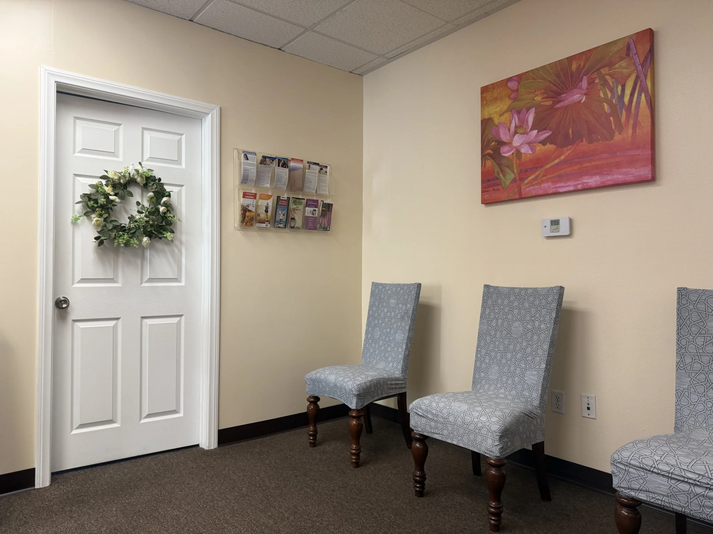 Interior of a waiting room with three patterned chairs, a door with a floral wreath, wall-mounted brochure rack, and colorful artwork on the wall.