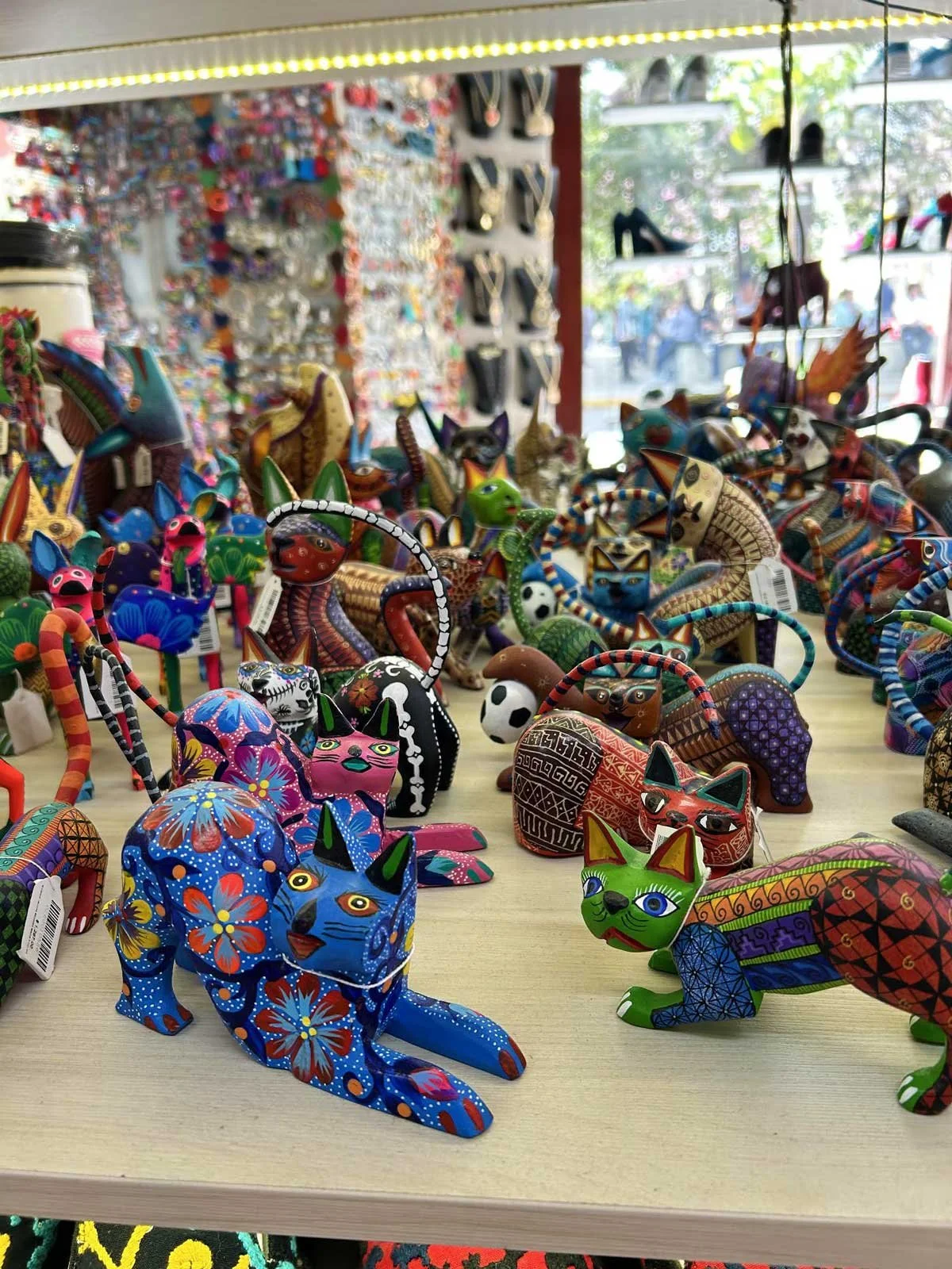 Colorful Mexican wooden cat figurines displayed on a table in a shop, with more in the background and outside.