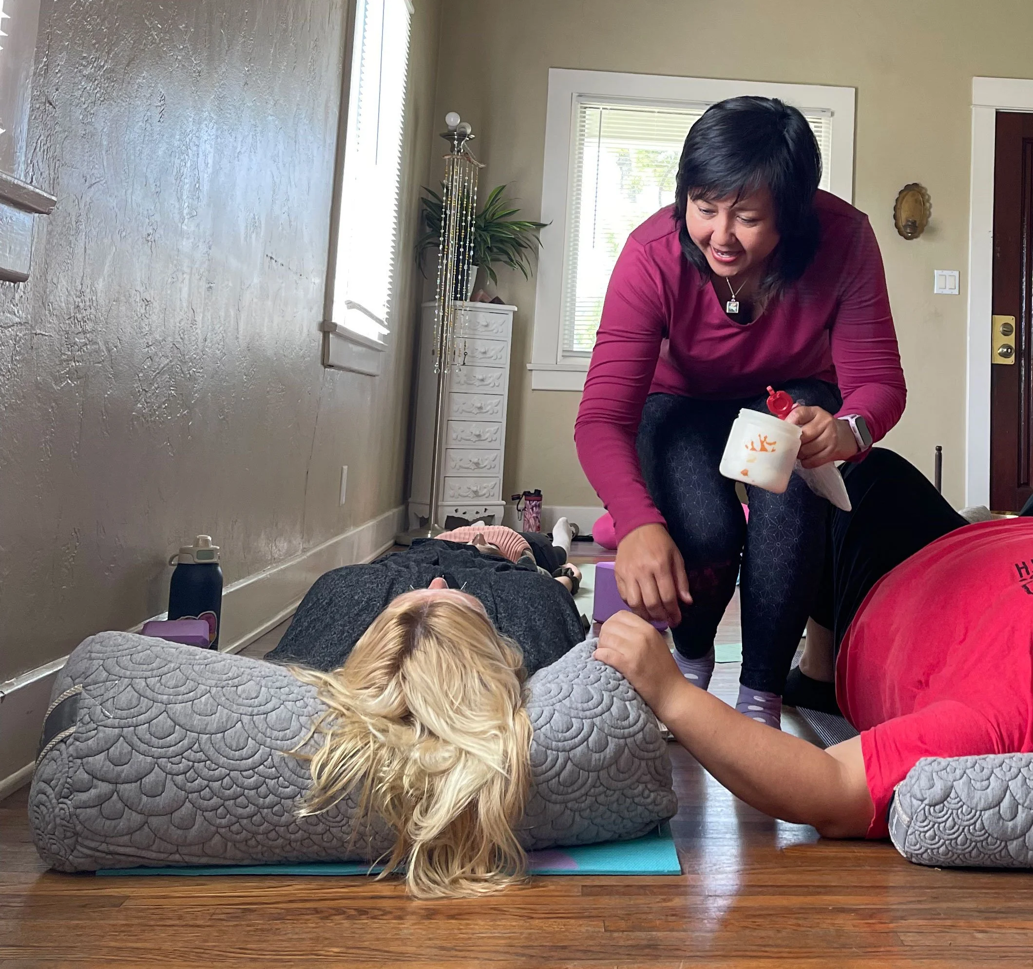 A woman assisting a person in a yoga exercise on the floor, with a woman in front of her lying face down on a mat, and a woman is smiling in upland, ca.