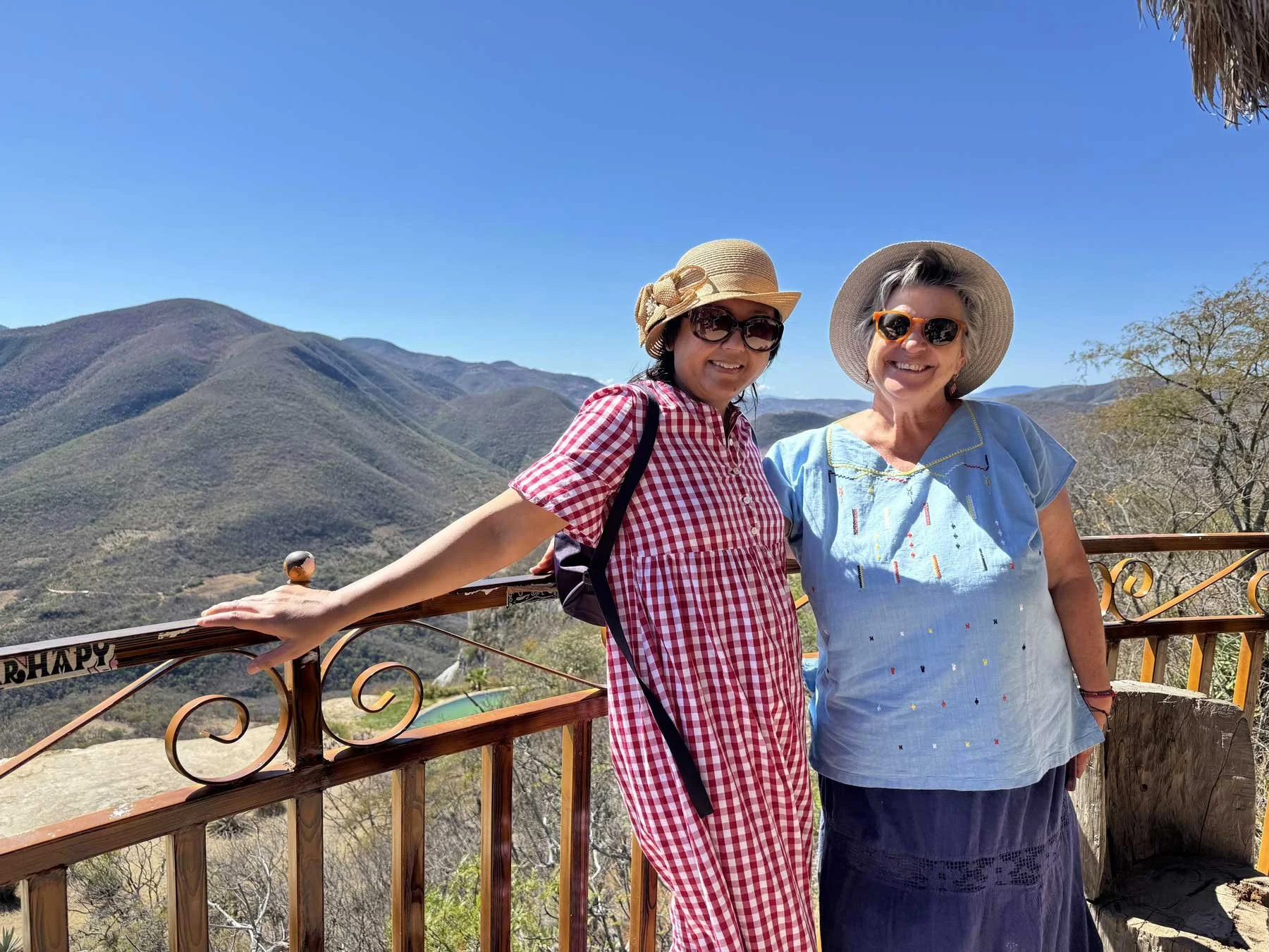 Two women smiling and wearing hats and sunglasses, standing on a wooden balcony overlooking a mountain landscape under a clear blue sky.