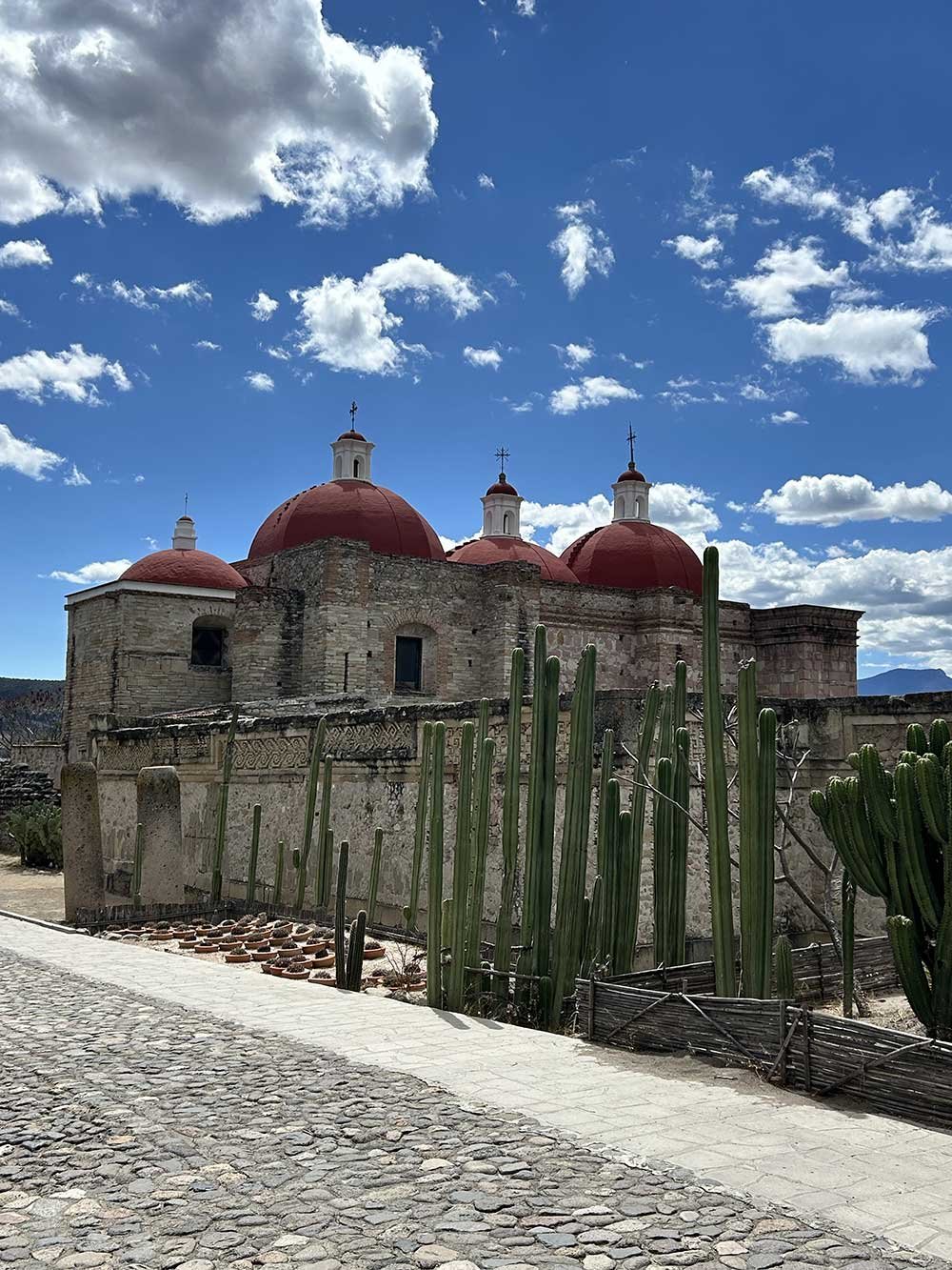 A historic church with multiple red domes, surrounded by tall cacti, under a partly cloudy blue sky.