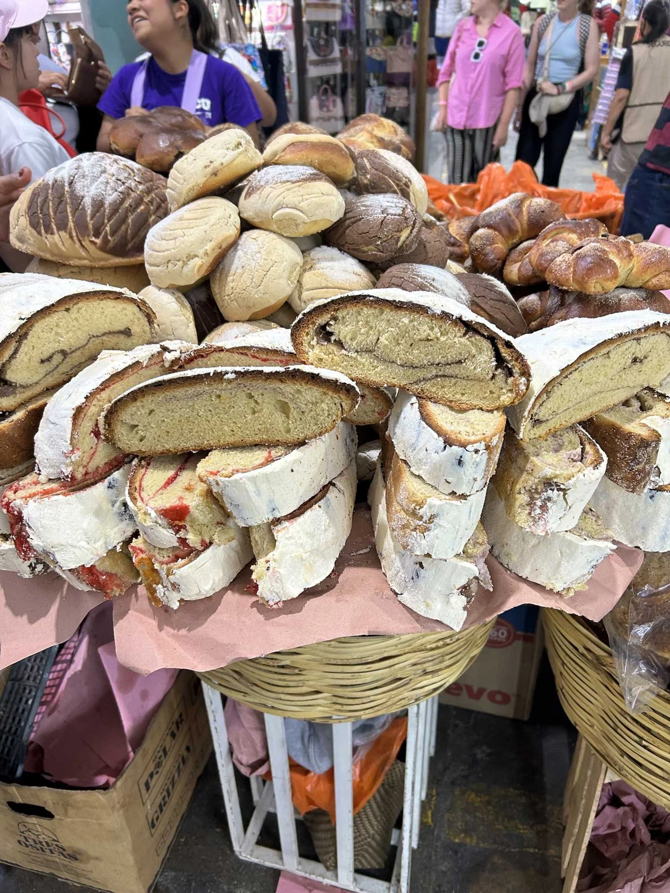 An assortment of baked goods including sliced rolls and whole pastries displayed on a basket at an outdoor market, with people shopping and browsing in the background.
