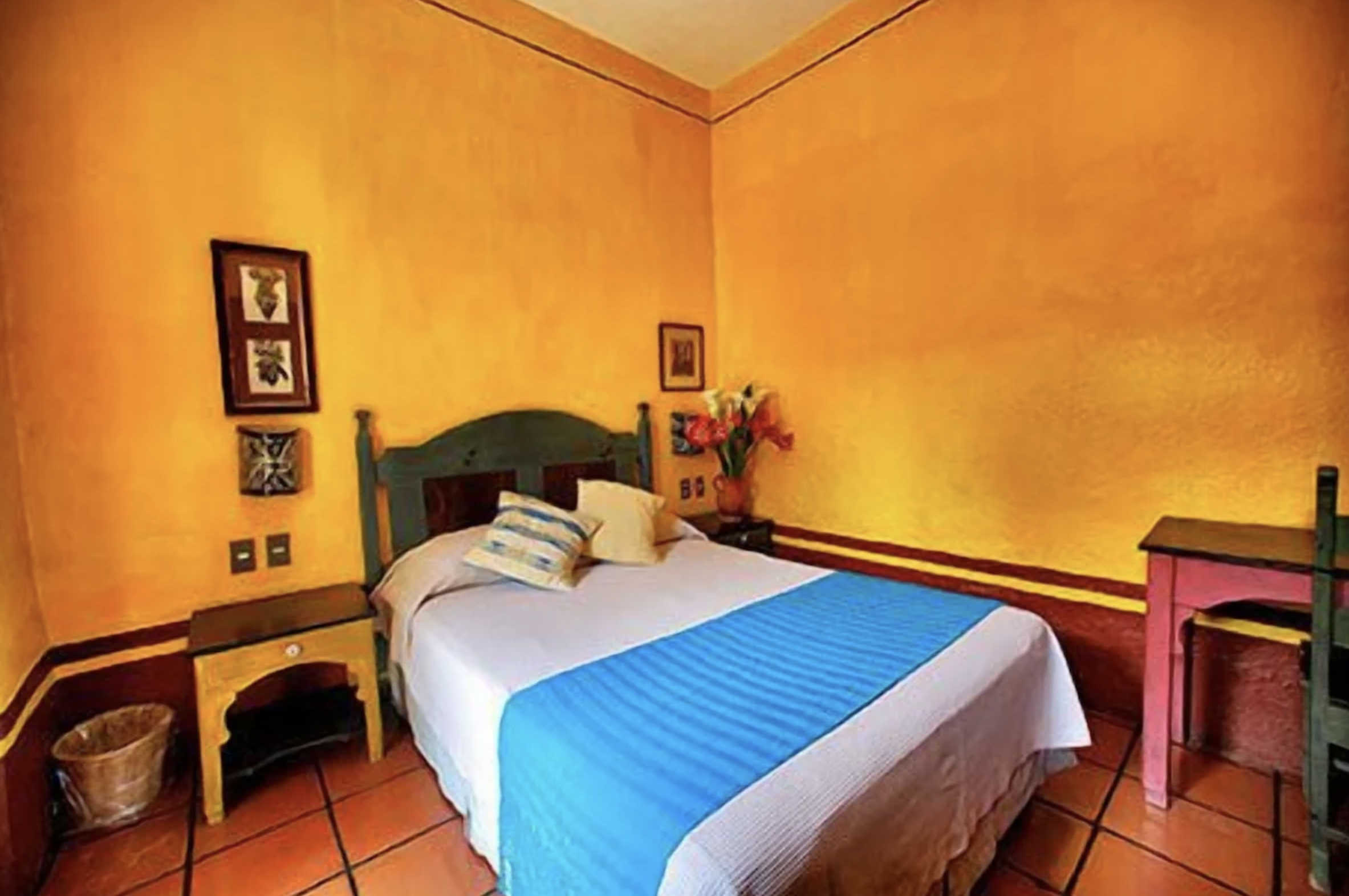 bedroom at Hotel Azucena in Oaxaca, Mexico. The room has yellow walls and white tile floors. There is a double bed in the center of the room, with a white bedspread and two pillows. On either side of the bed there is a nightstand. The nightstand on t