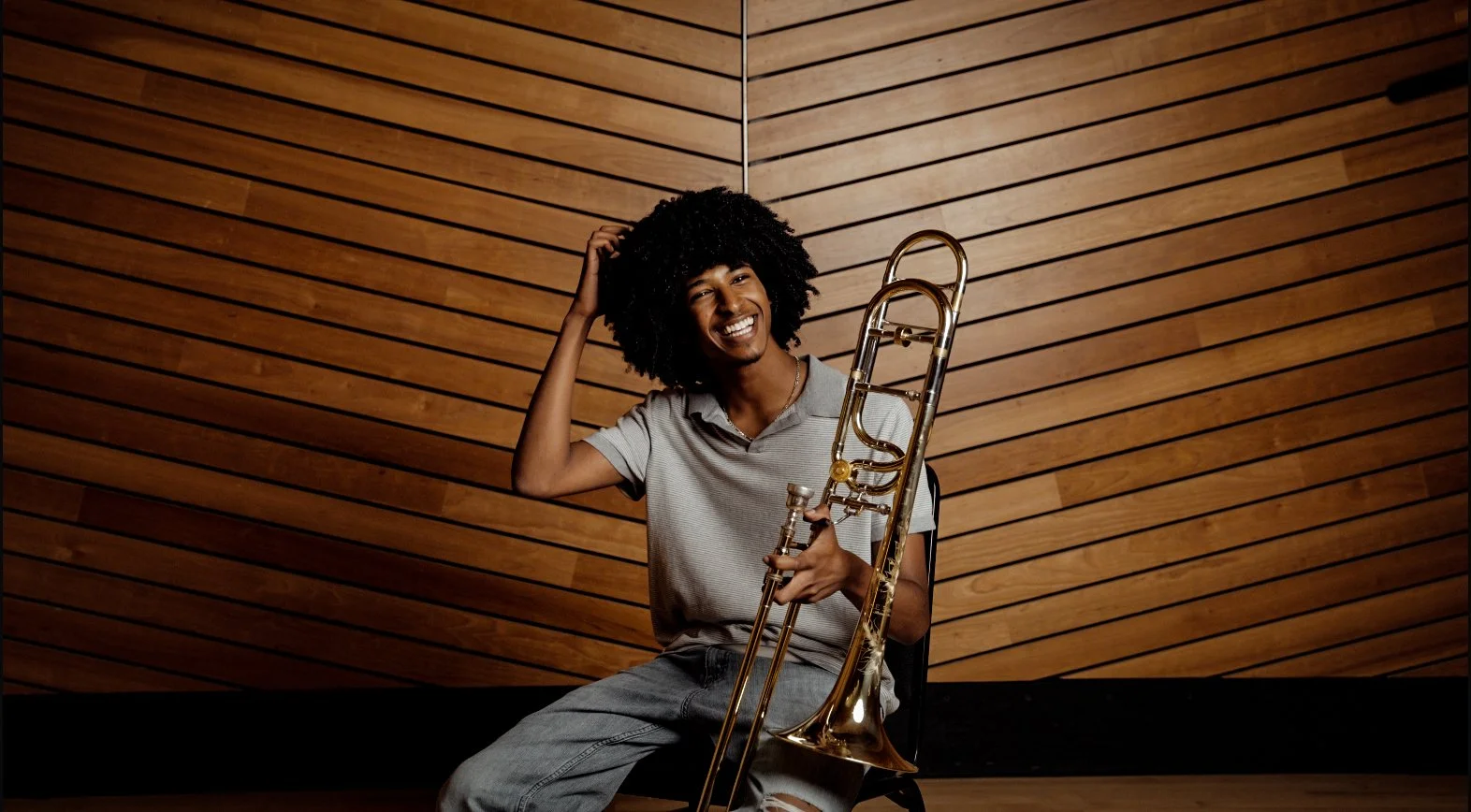 A young person with curly hair sitting in a recording studio, smiling, holding a brass trombone, with a wooden paneled wall background.