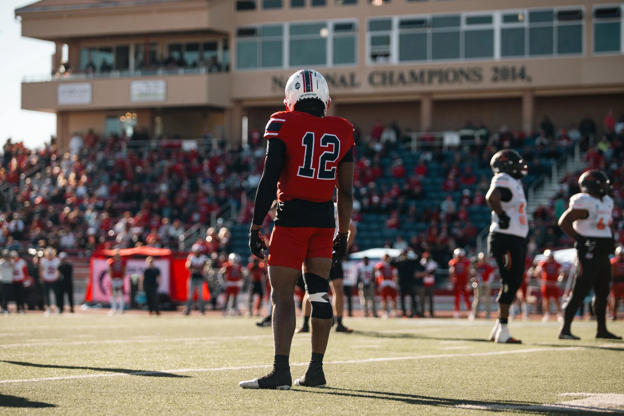 Football player wearing a red jersey with the number 12, standing on the field during a game, with spectators in the stands and other players nearby.