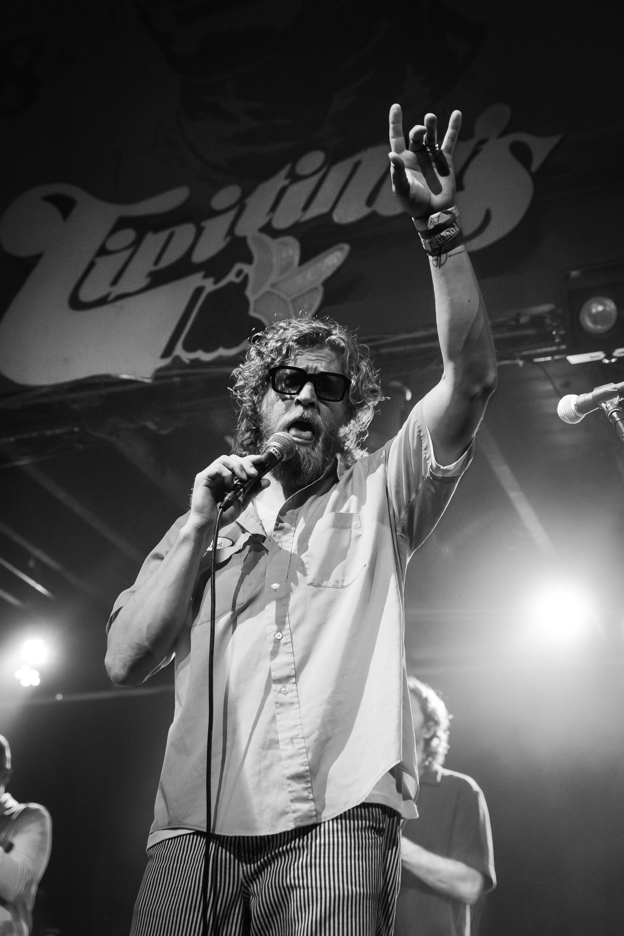 A man with curly hair and a beard wearing sunglasses, a short-sleeved button-up shirt, and striped pants, holding a microphone and raising one arm in front of a stage with a 'Lipton's Ice Tea' sign.