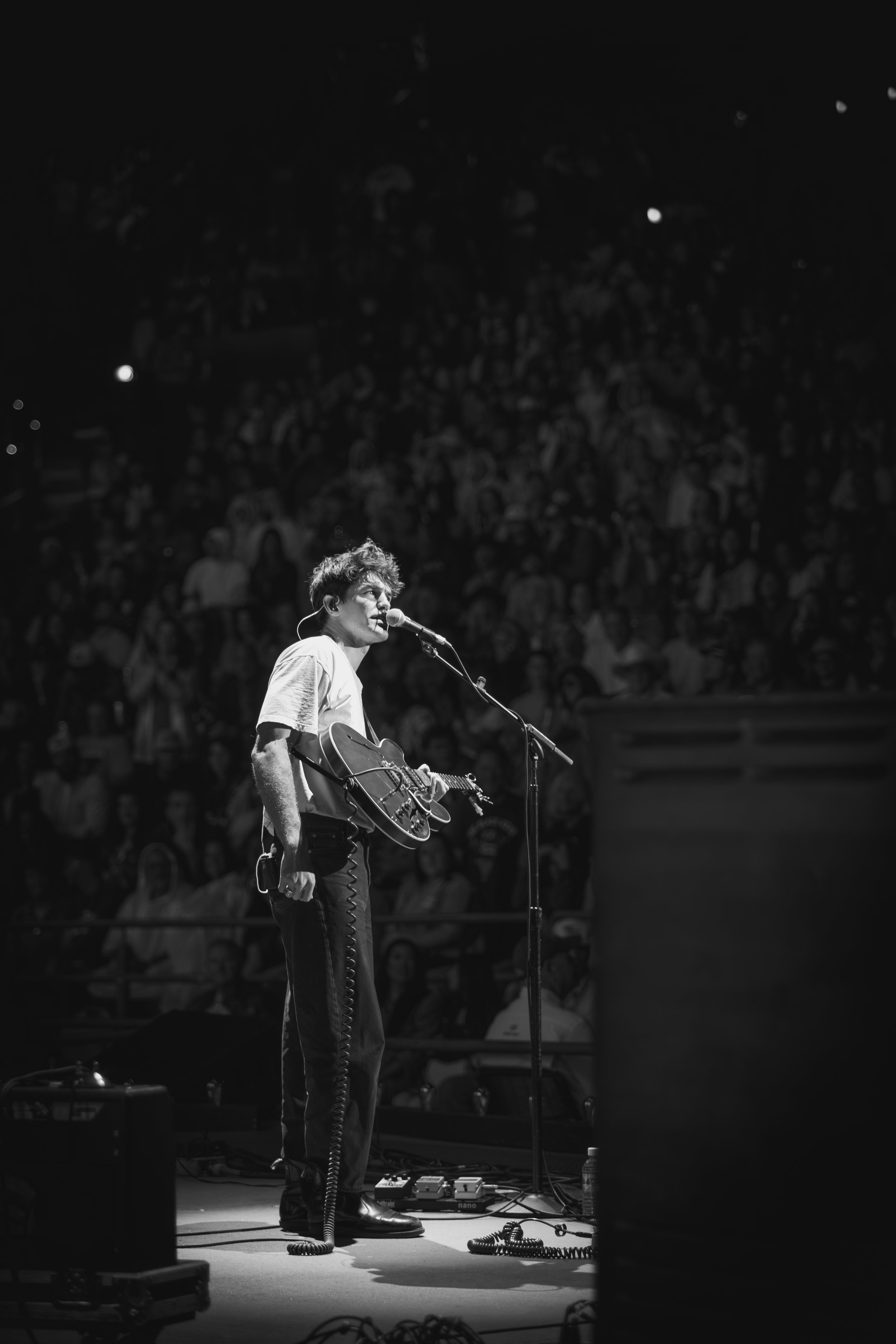 A musician performing on stage with an acoustic guitar and microphone, in front of a large audience, in black and white.