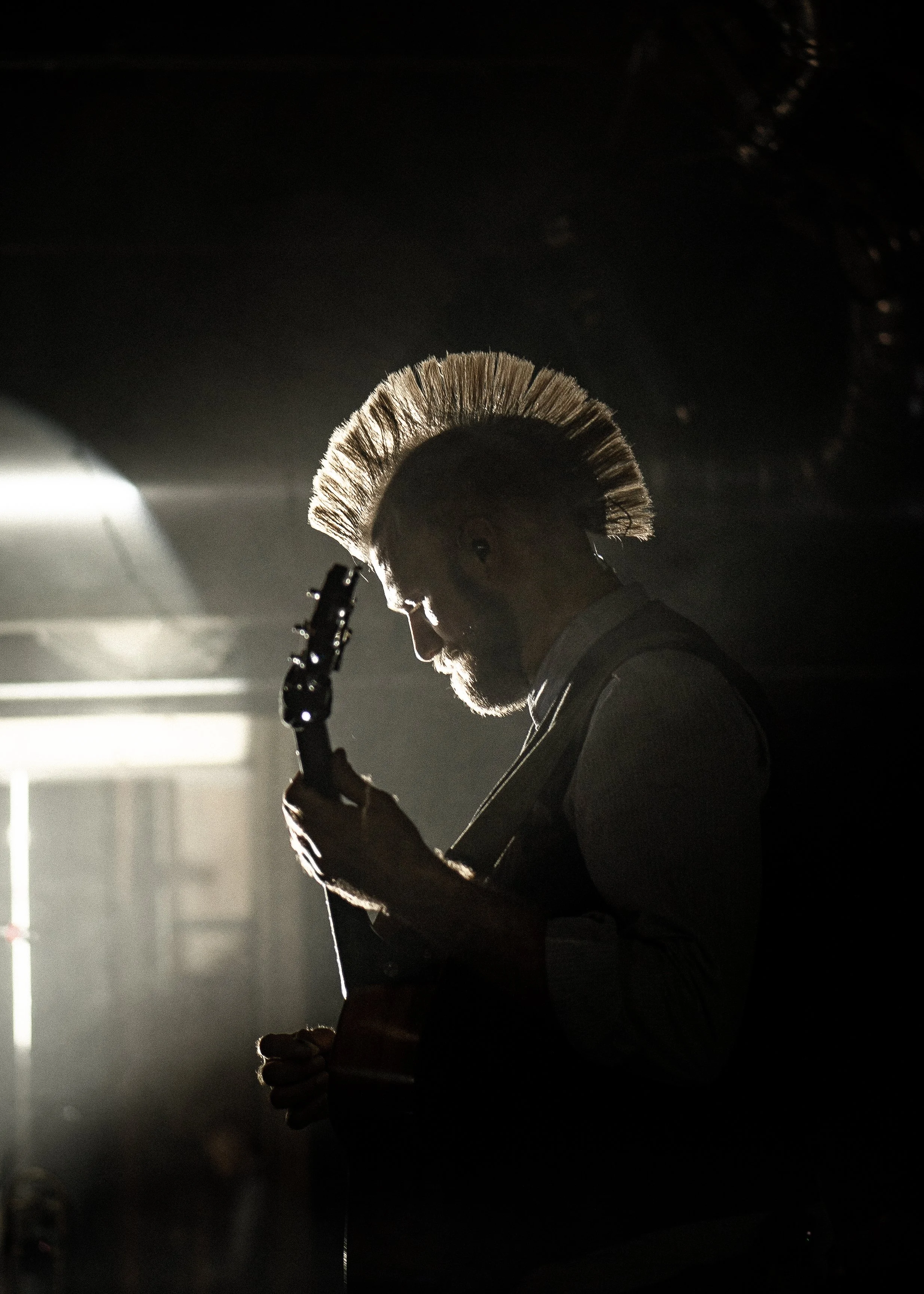 A man with a mohawk playing an electric guitar on stage, backlit with a bright light creating a silhouette effect.