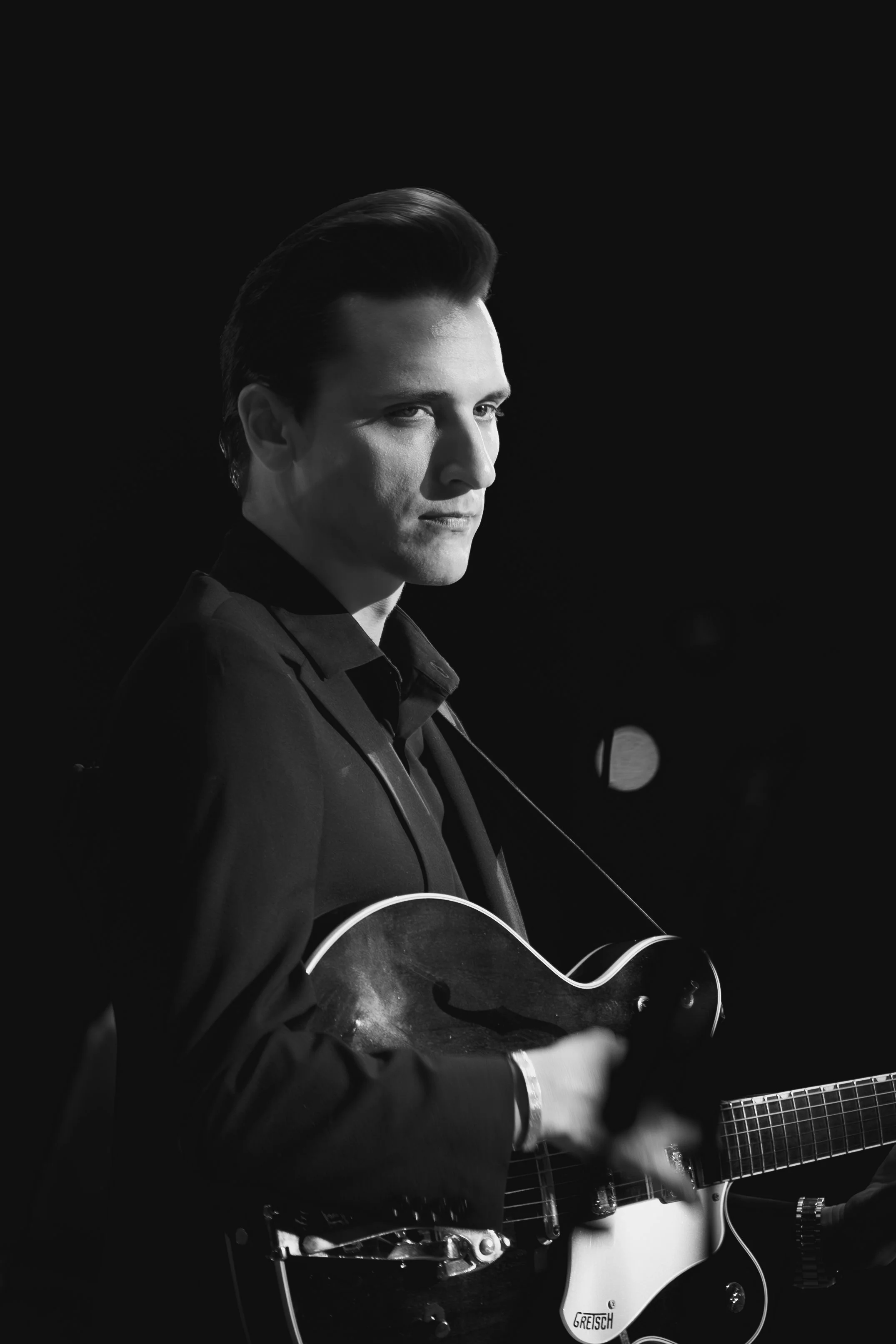 A black and white photo of a man with slicked-back hair playing an electric guitar, wearing a dark suit, against a dark background.
