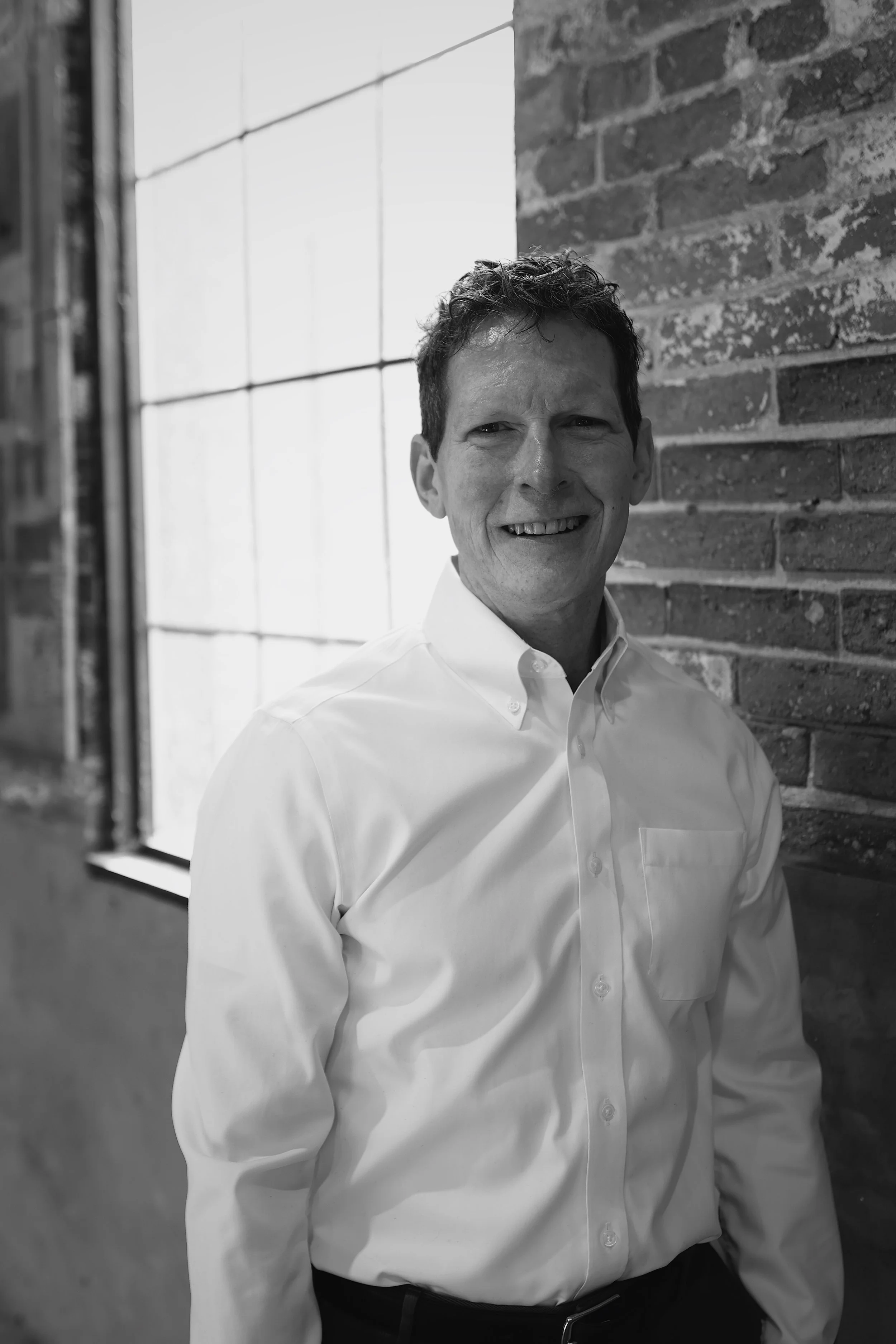 Man in a black shirt and black cap standing outside near a brick wall and glass-paneled door with greenery.