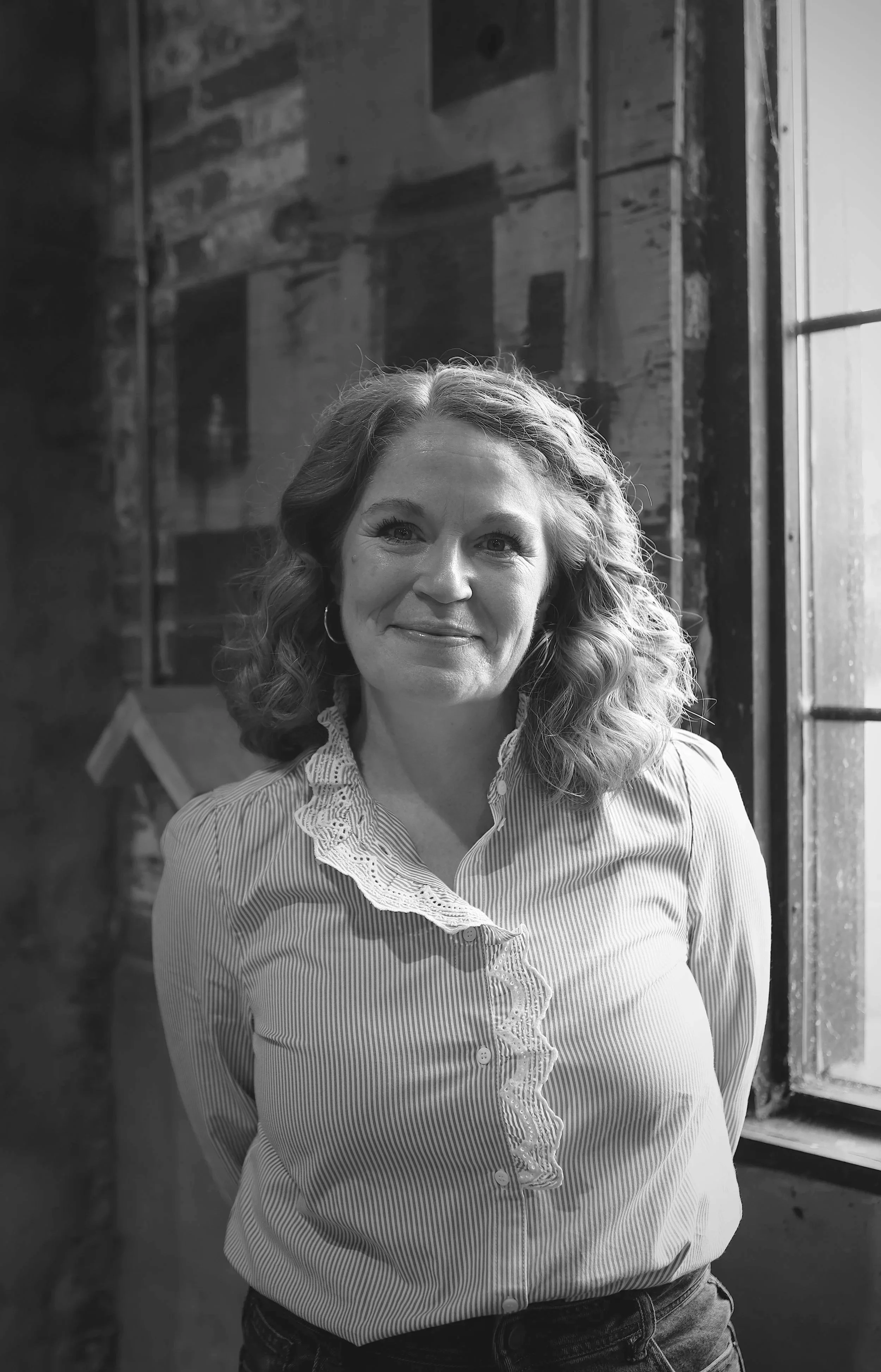 Smiling woman in a gray t-shirt and jeans standing outside near green foliage and a brick wall.