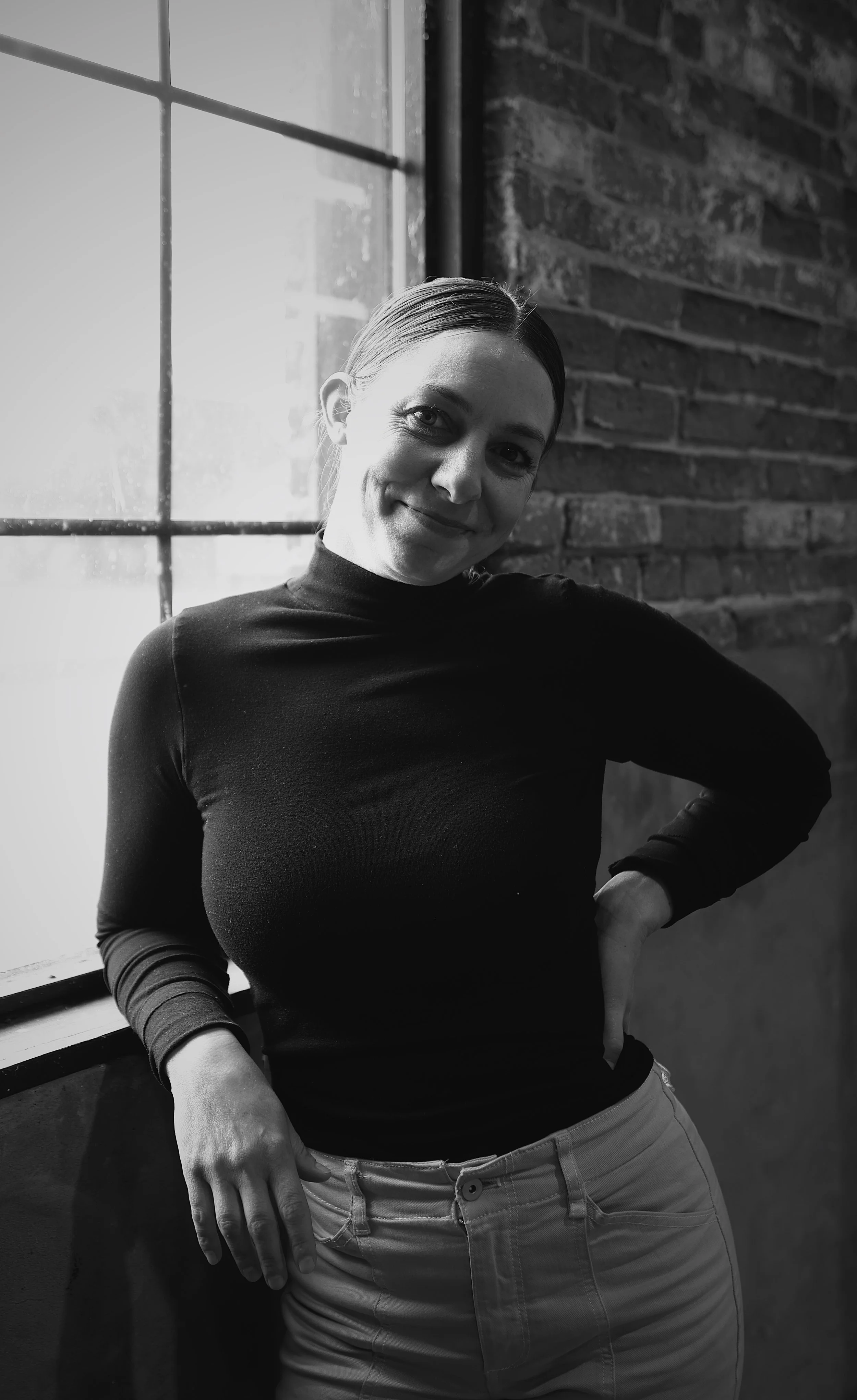 Person with short brown hair smiling, wearing a light-colored shirt and jeans, standing outdoors near a brick wall and greenery.