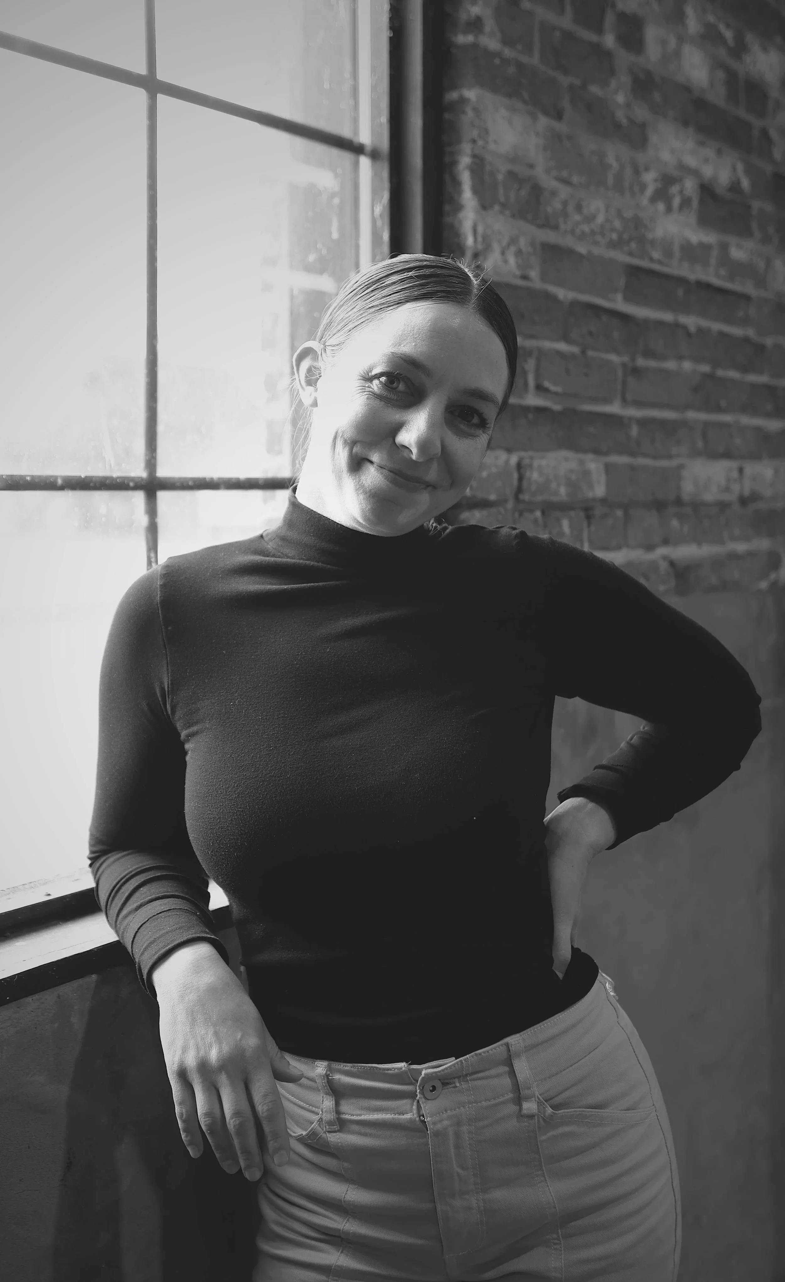 Person with short brown hair smiling, wearing a light-colored shirt and jeans, standing outdoors near a brick wall and greenery.