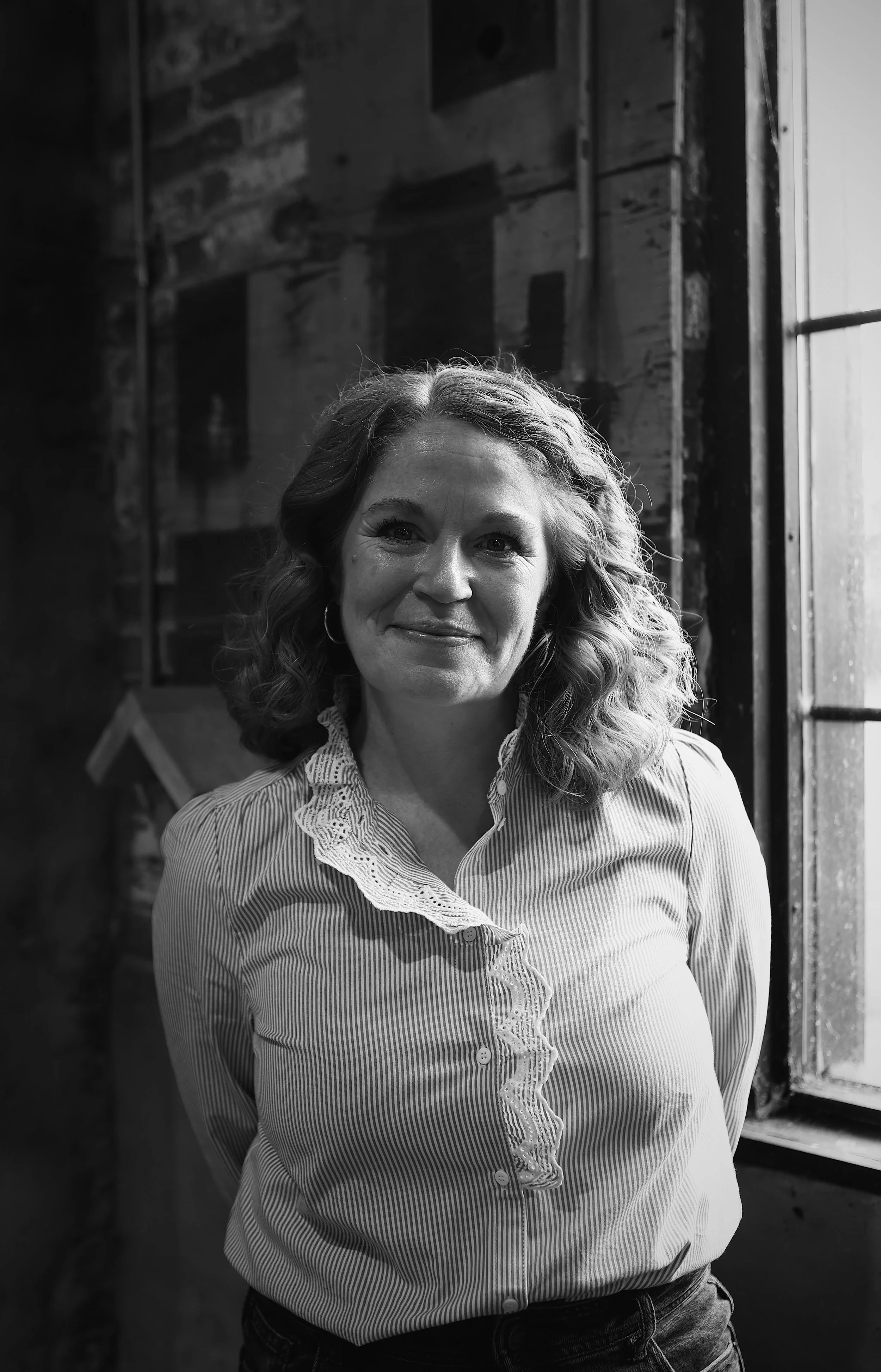 Smiling woman in a gray t-shirt and jeans standing outside near green foliage and a brick wall.