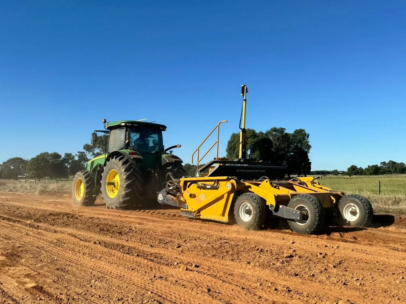 A green tractor pulling a yellow land roller on a dirt field under a clear blue sky.