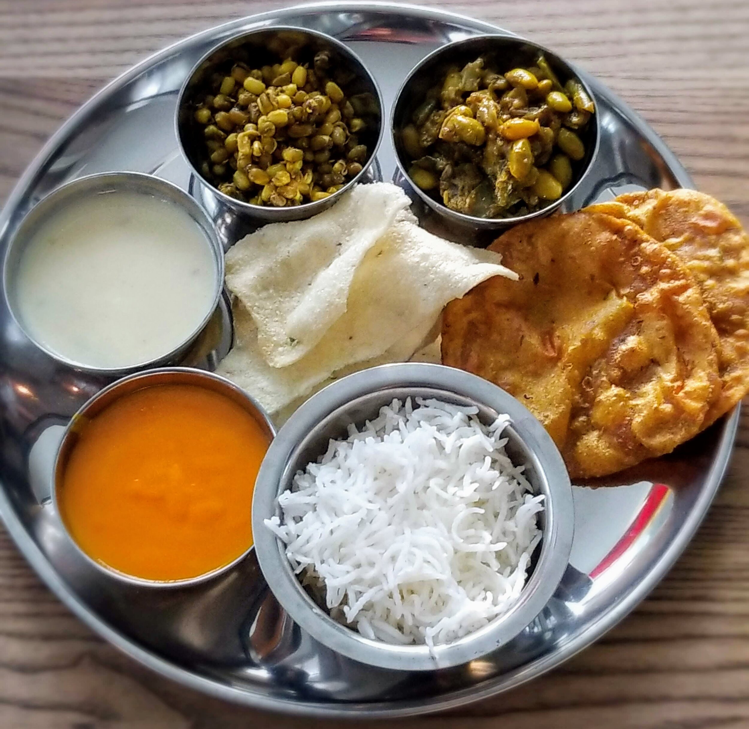 A thali is an assortment of small dishes served as a meal that differs from region to region. My family is from Gujarat, a state in west India known for its vegetarian dishes, so many of the recipes you find on here will tend to be the Gujarati version of Indian dishes. The typical components of a Gujarati thali are rice, a bread item like puris, a few vegetable dishes, lentil gravies, and a sweet. Here we have the curried mung beans along with Basmati rice and puris, whose recipes you can find in the recipe box. We’re going to work our way around the thali, learning about kadhi, the white gravy made from chickpeas; papdi, a crunchy snack made from rice flour; papdi saak, another Indian vegetable dish.; and sweet and orange mango rus.