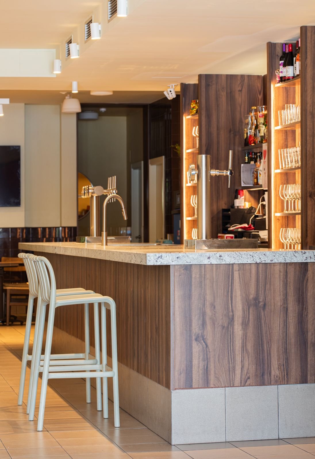 Bar con encimera de piedra, estanterías de madera con botellas y vasos, y sillas blancas en primer plano.