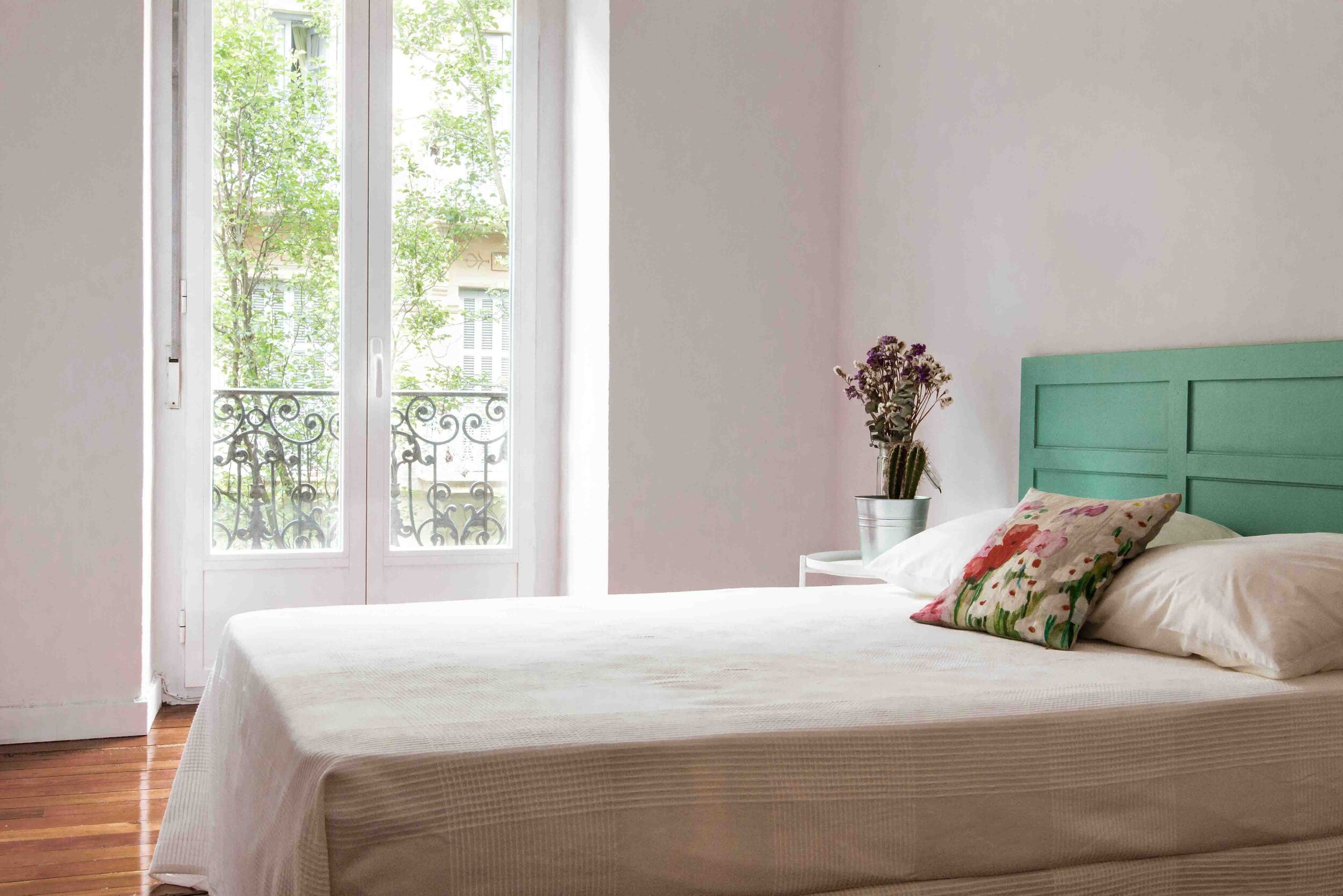 Habitación con cama con cabecera verde, almohada decorativa con flores, ventana con vista a balcón y árboles, piso de madera, paredes blancas, planta en maceta al lado de la cama.