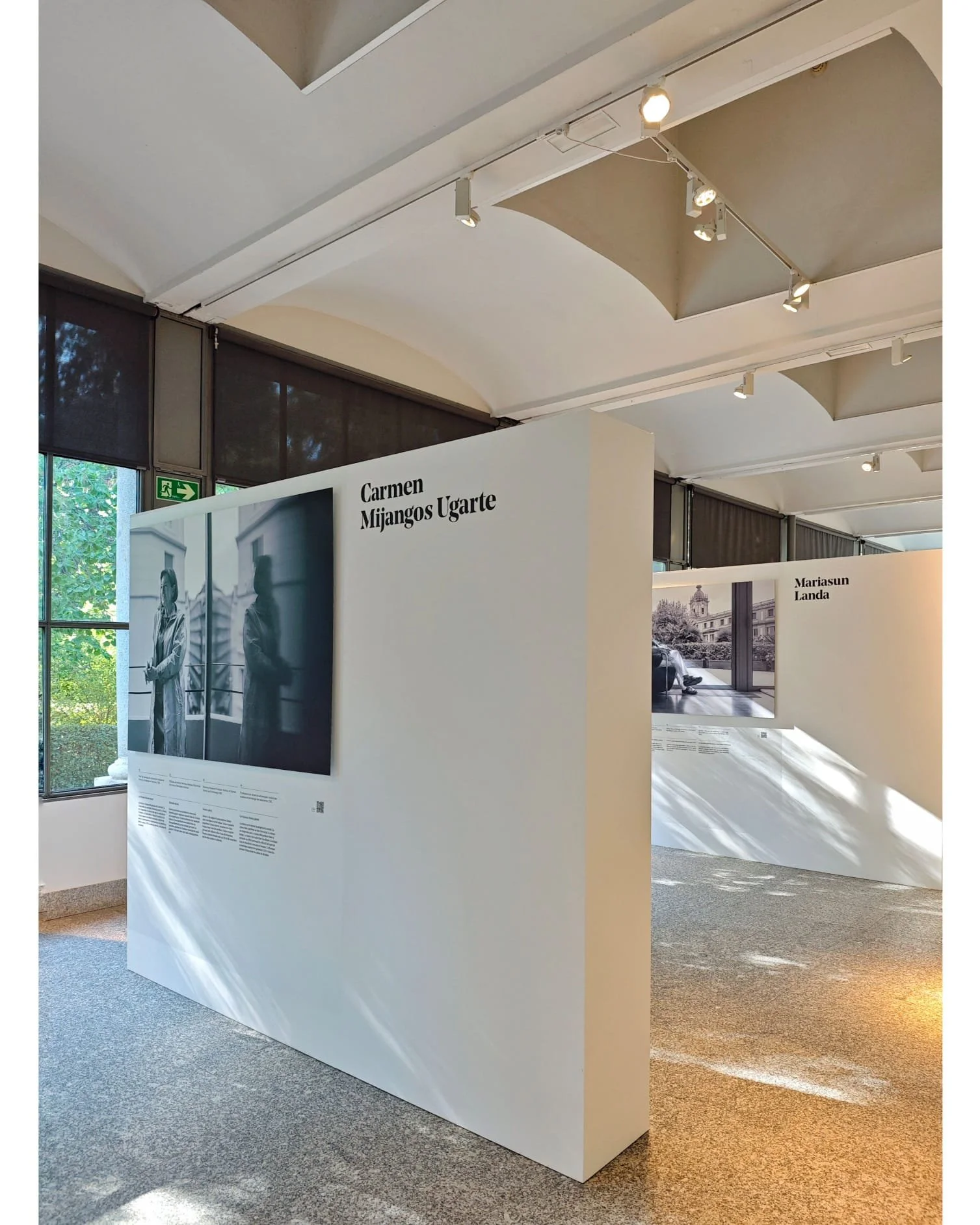 Exposición en un museo con paneles blancos que muestran fotografías en blanco y negro y textos explicativos, con nombres de artistas como Carmen Mijangos Ugarte y Mariasun Landa, y figuras humanas en las fotografías.