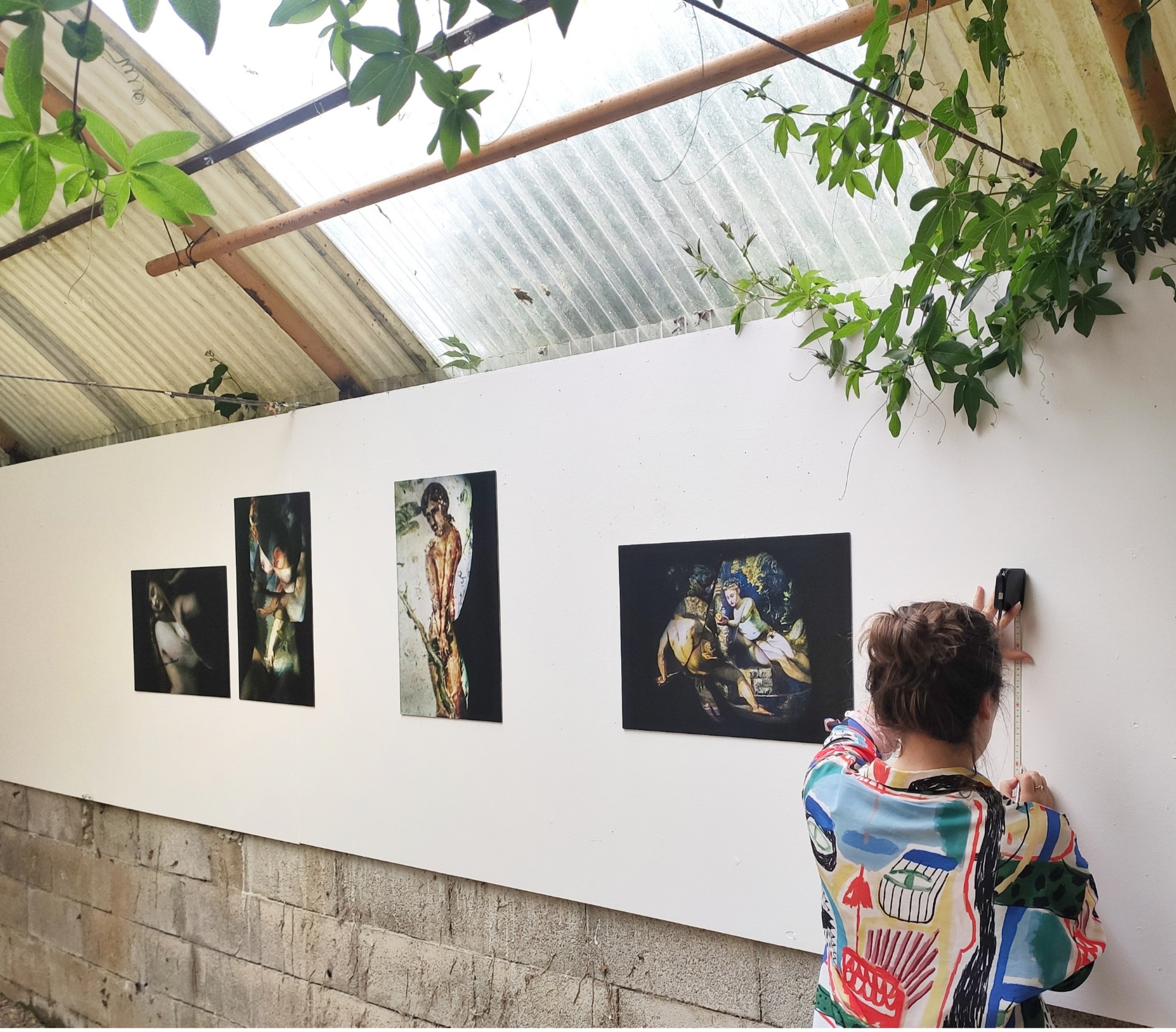 Persona observando y midiendo una obra de arte en una galería con paredes blancas y varias pinturas expuestas, techo de metal y plantas colgantes.