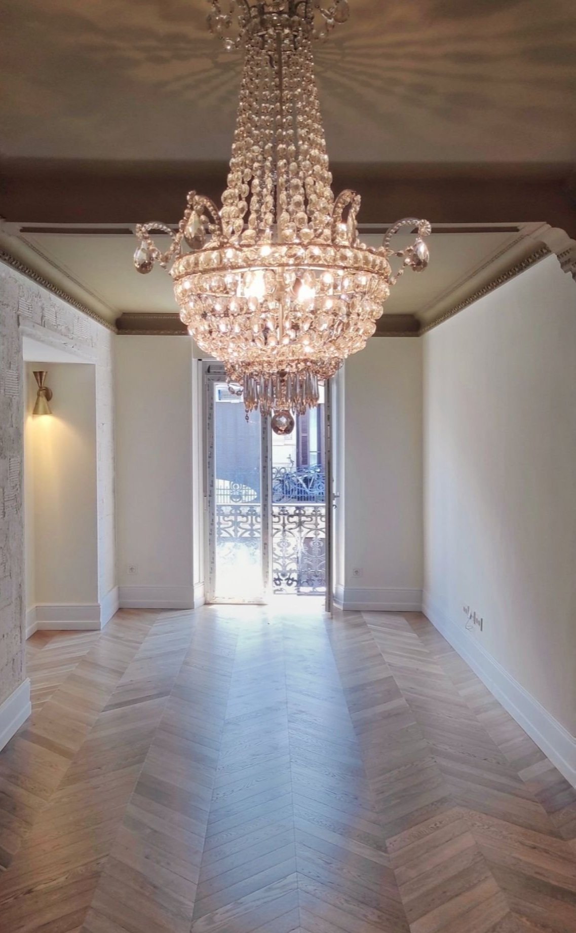 Sala elegante con un gran candelabro de cristal y piso de madera en damero, con balcón en el fondo.