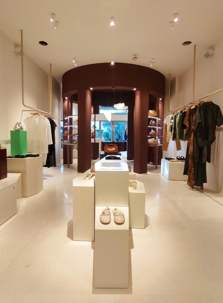 Interior de tienda de moda y accesorios con ropa en exhibición y estanterías de zapatos y bolsos.