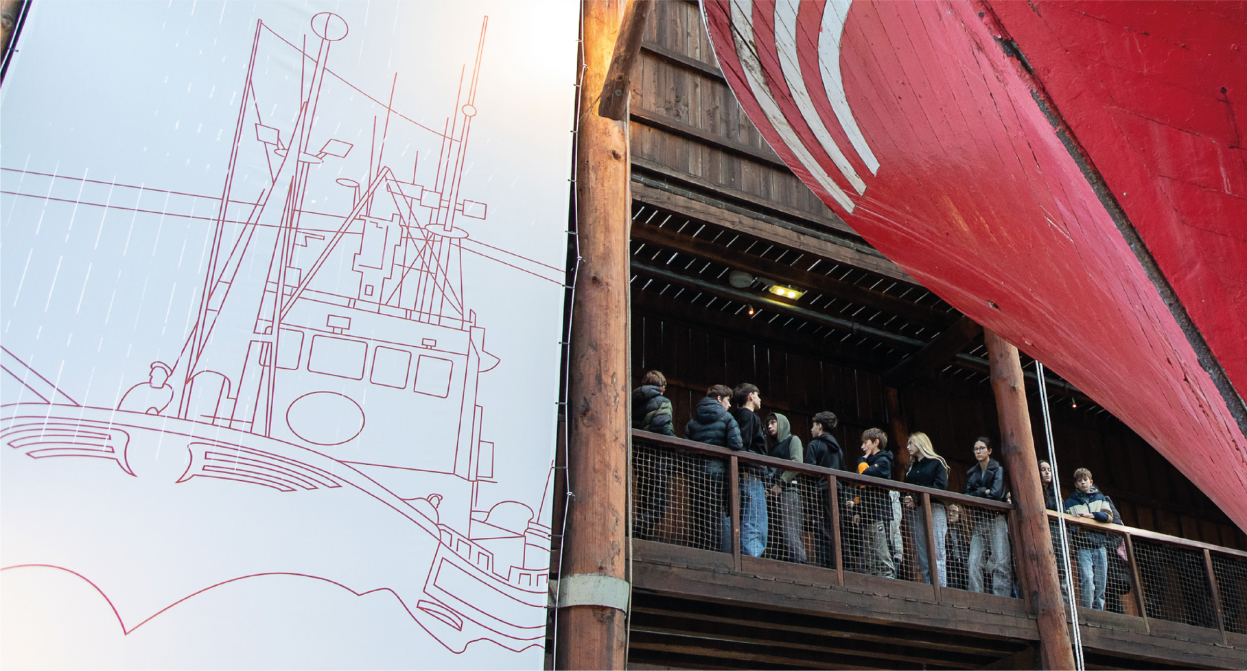 Grupo de personas en una fila dentro de un edificio de madera, junto a un barco grande, en un museo o exhibición, con un esquema de un barco en la pared