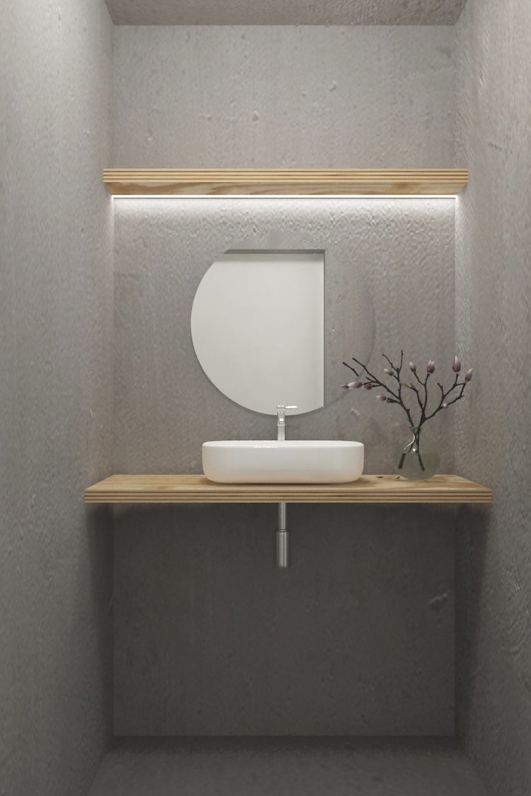 Lavabo rectangular blanco sobre una repisa de madera natural, con un espejo redondo sobre la repisa y un jarrón con ramas y flores en la esquina derecha, en una pared de textura gris.