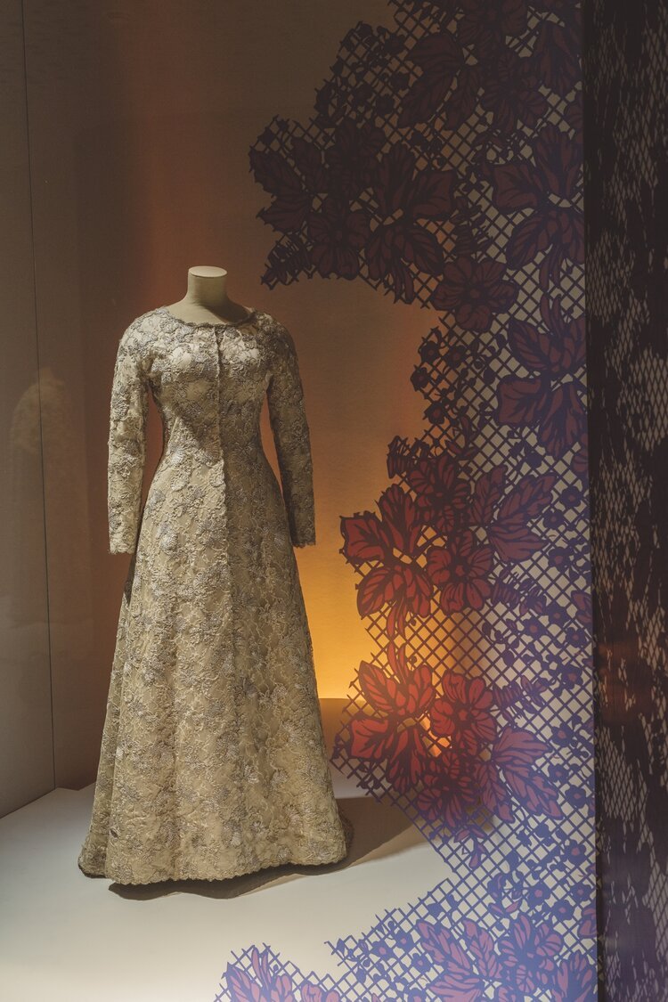 Vestido largo de encaje y manga larga en exhibición, con fondo de iluminación cálida y decoración de flores y patrones de malla en tonos rojos y morados.