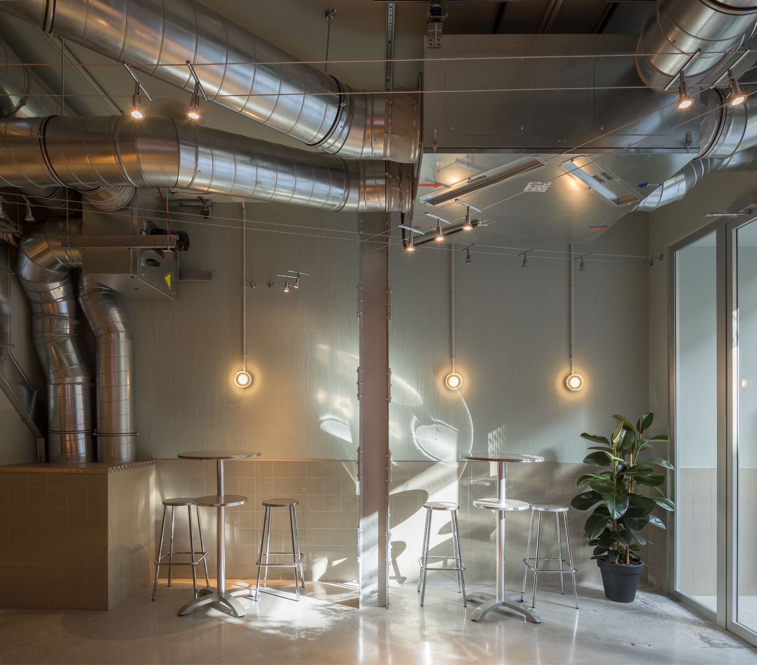 Interior de un café con mesas y bancos de metal, planta y ventilaciones expuestas en el techo.