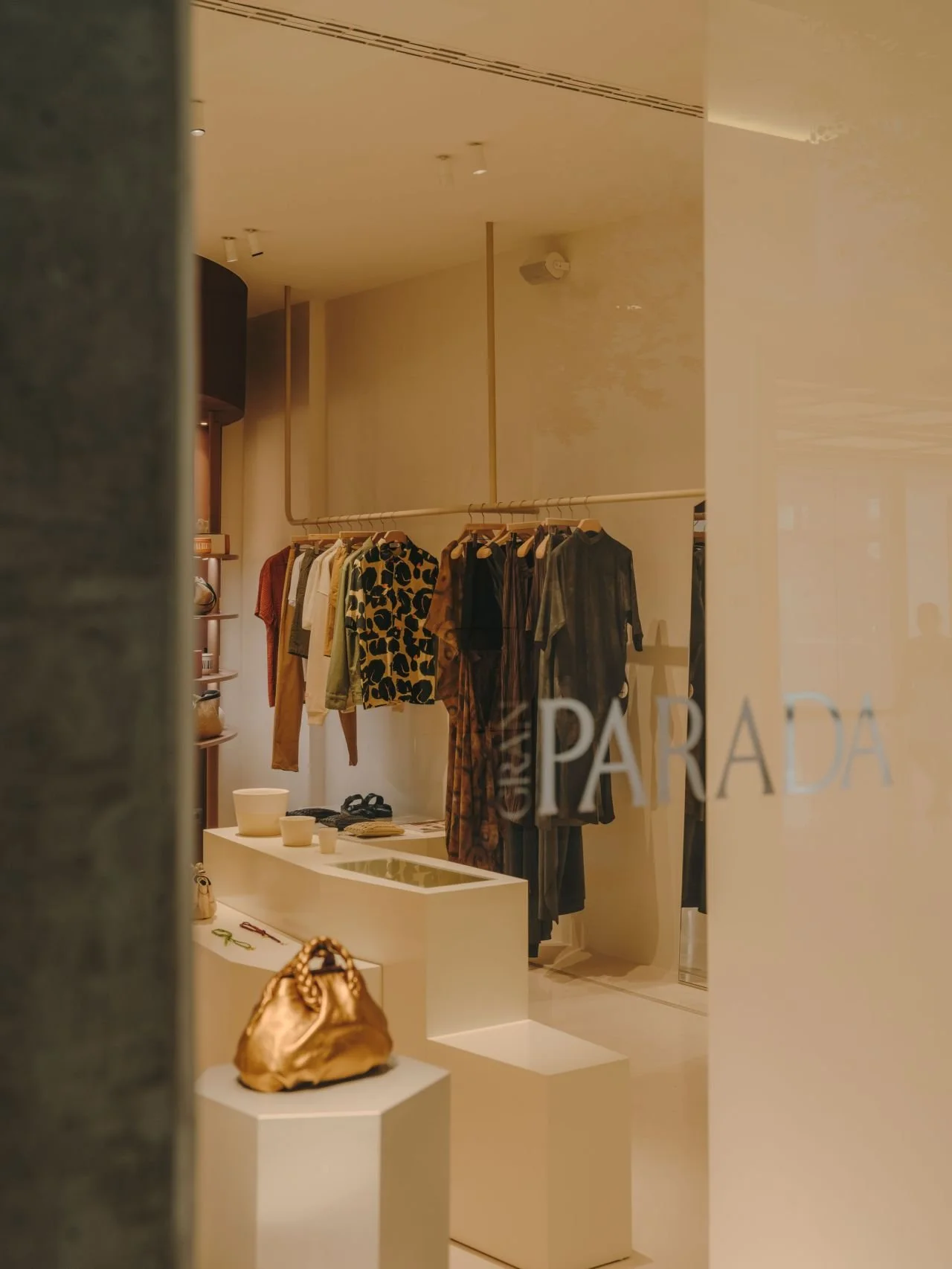 Vista del interior de una tienda de ropa con prendas en perchas y accesorios en mesas, vista a través de un cristal con la palabra 'PARADA' escrita.