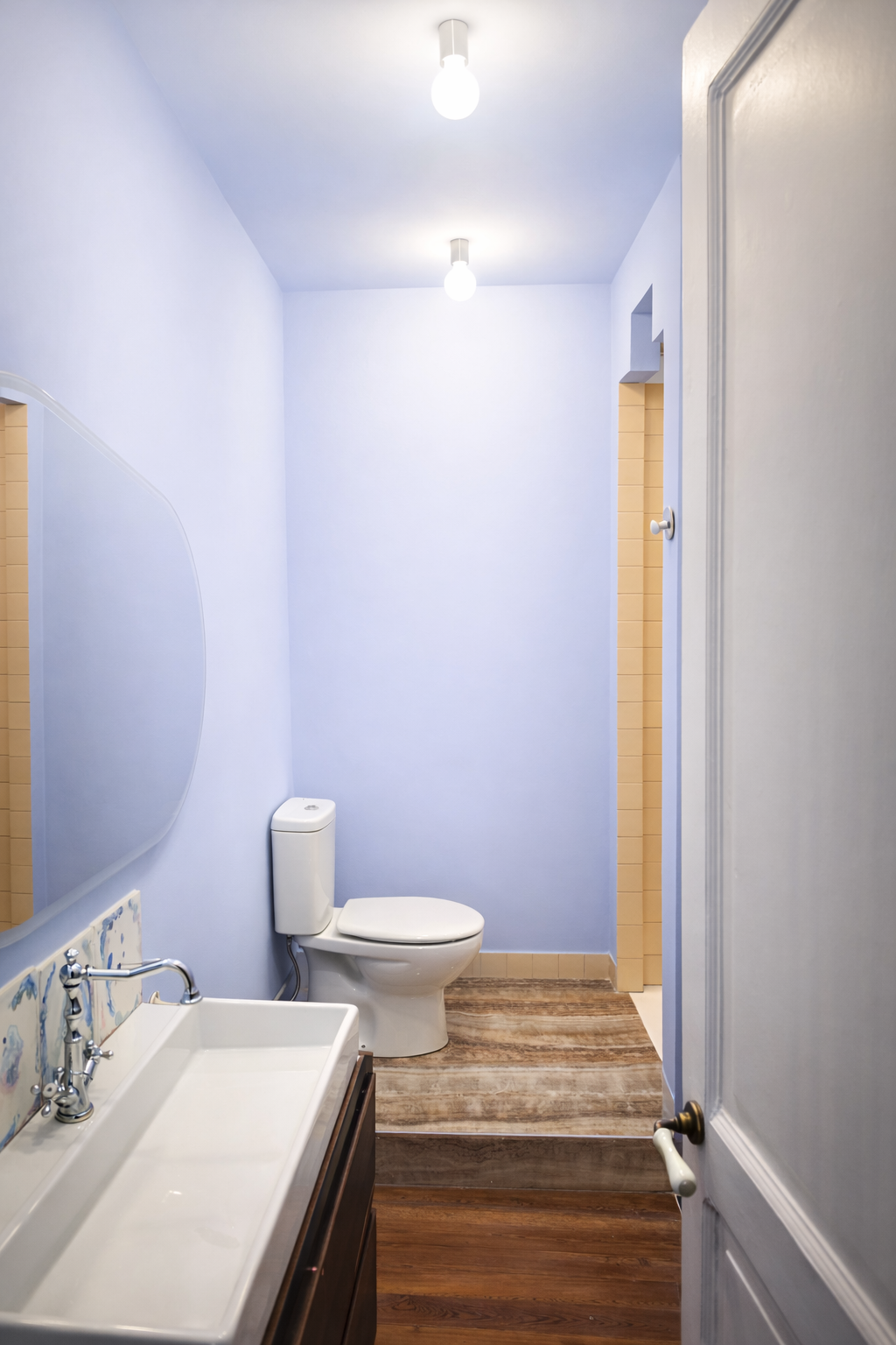 Baño con paredes azules, espejo, lavabo con mueble, inodoro, y piso de madera.
