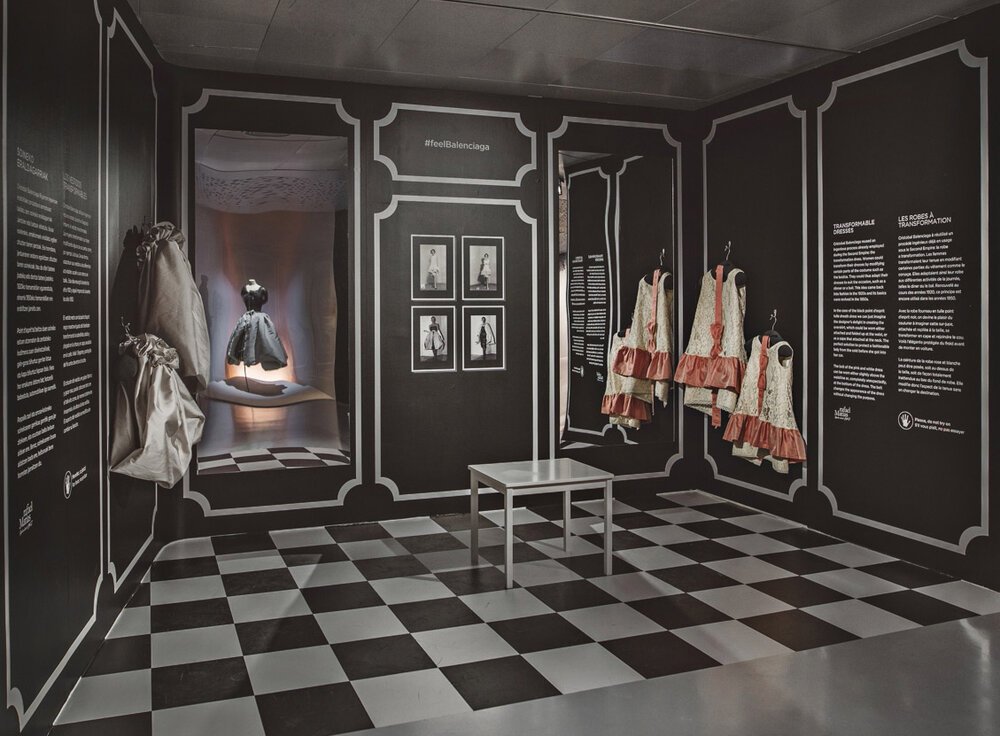 Una exhibición en un museo de moda que muestra ropa histórica y fotografías en un fondo negro con diseños blancos. En la parte izquierda hay vestuarios y accesorios, en el centro hay una publicación con imágenes, y en la derecha hay vestidos con deta