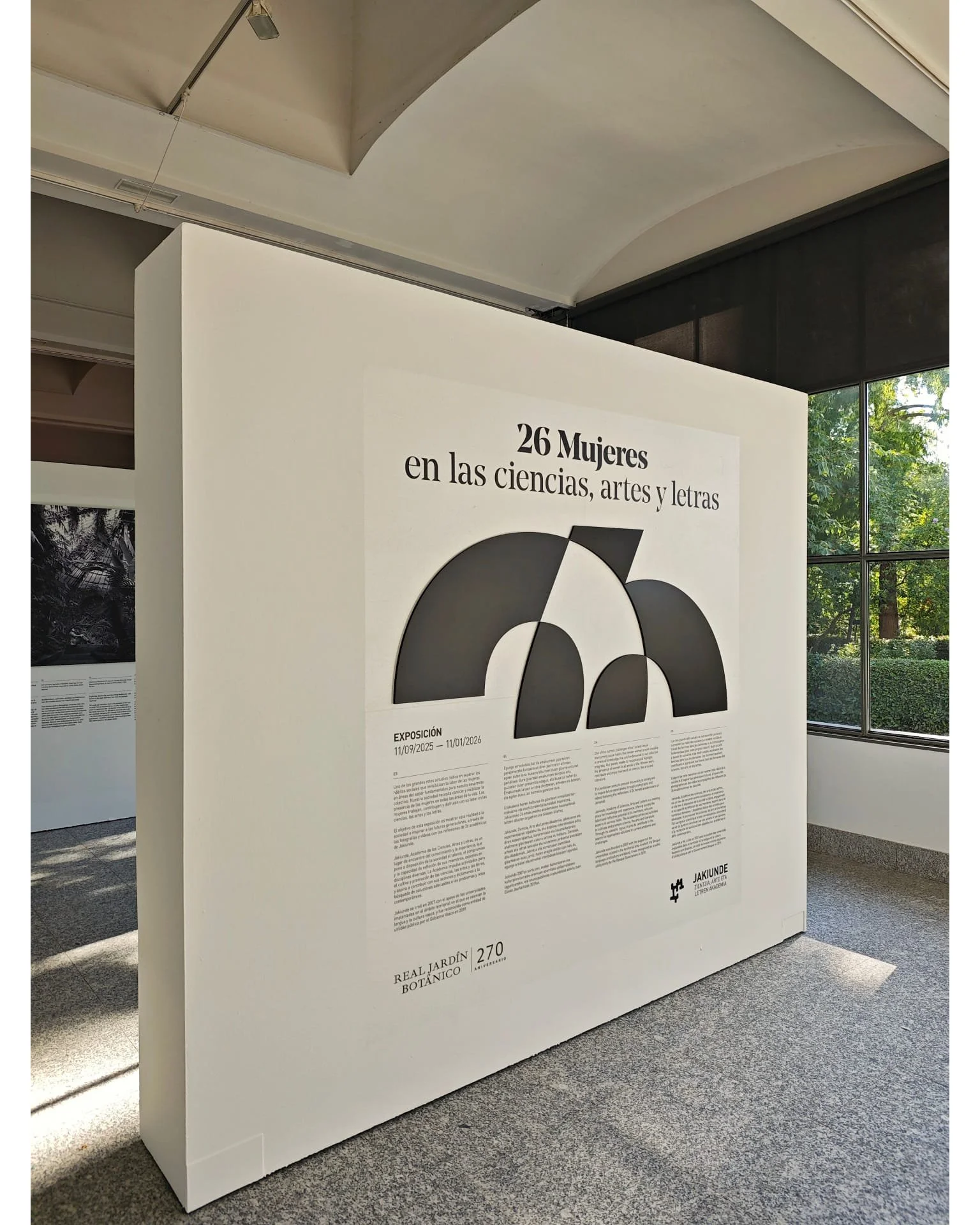 Cartel de exposición que muestra que hay 26 mujeres en ciencias, artes y letras. Incluye texto en español y gráficos geométricos en negro y blanco, con detalles de fecha y lugar de la exposición.