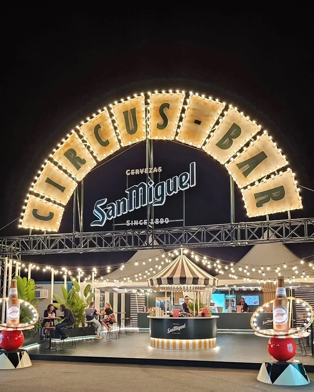 Escaparate de un establecimiento de cervezas San Miguel con iluminación decorativa, mesas, un mostrador y clientes en la terraza nocturna.