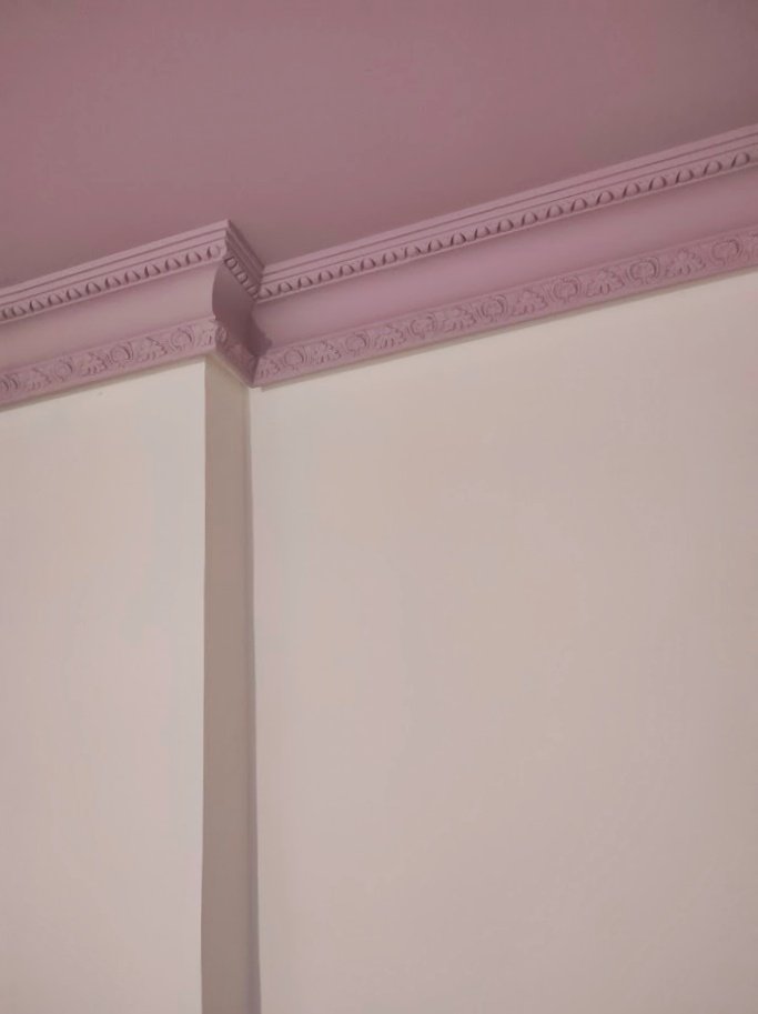 Detalle de la esquina de un techo con moldura decorativa en color lila y paredes blancas.
