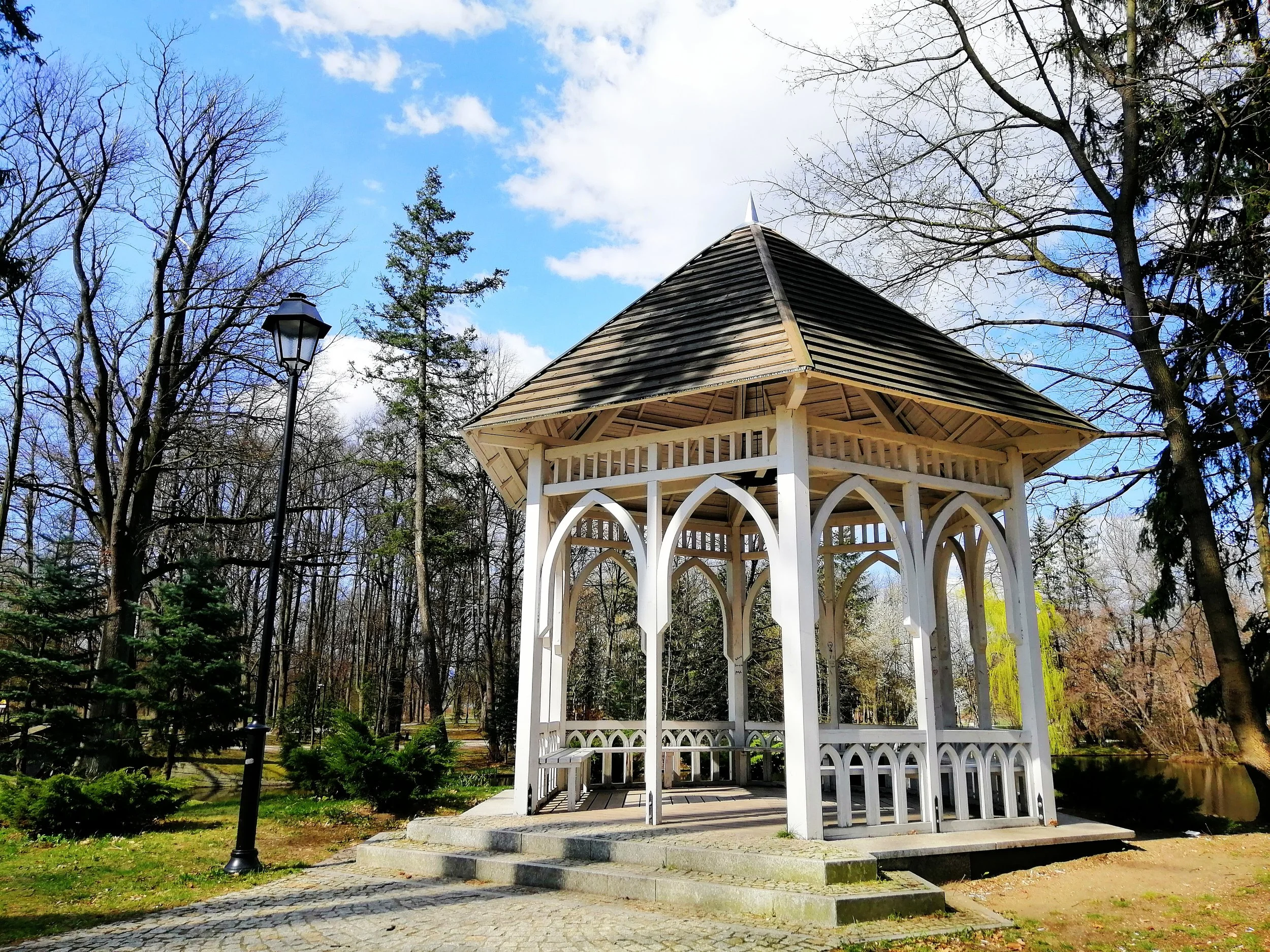 White gazebo in a park with trees, a cobblestone path, and a lamppost under a partly cloudy sky.